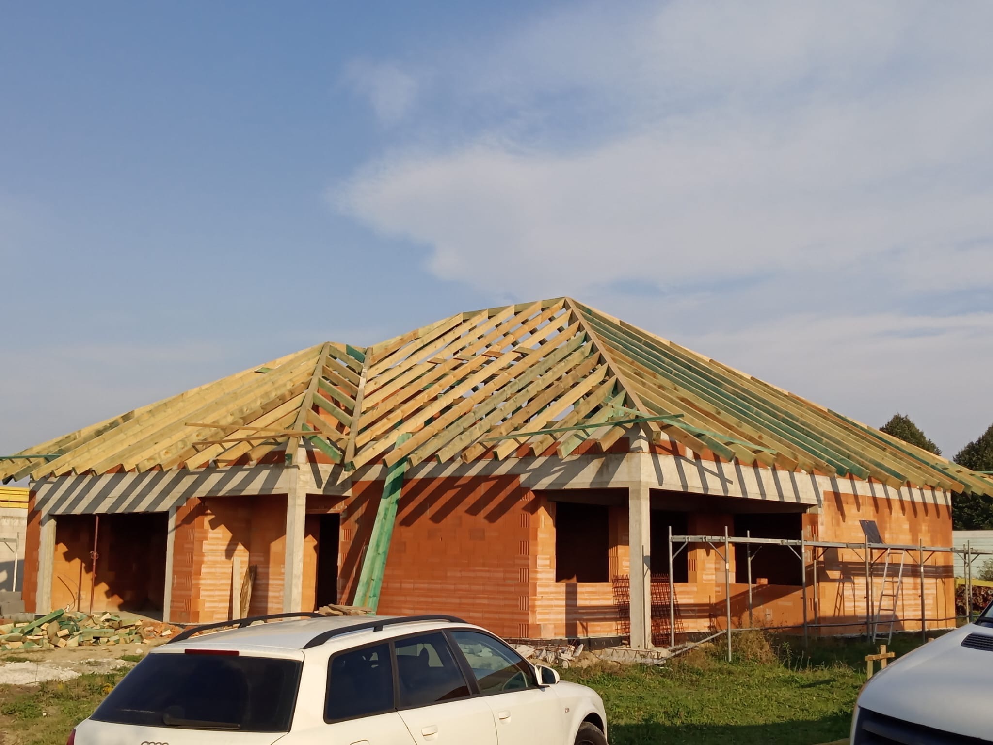 Budowa domu jednorodzinnego z czerwonej cegły. Widoczne ściany i drewniana konstrukcja dachu. Na pierwszym planie zaparkowane samochody osobowe.