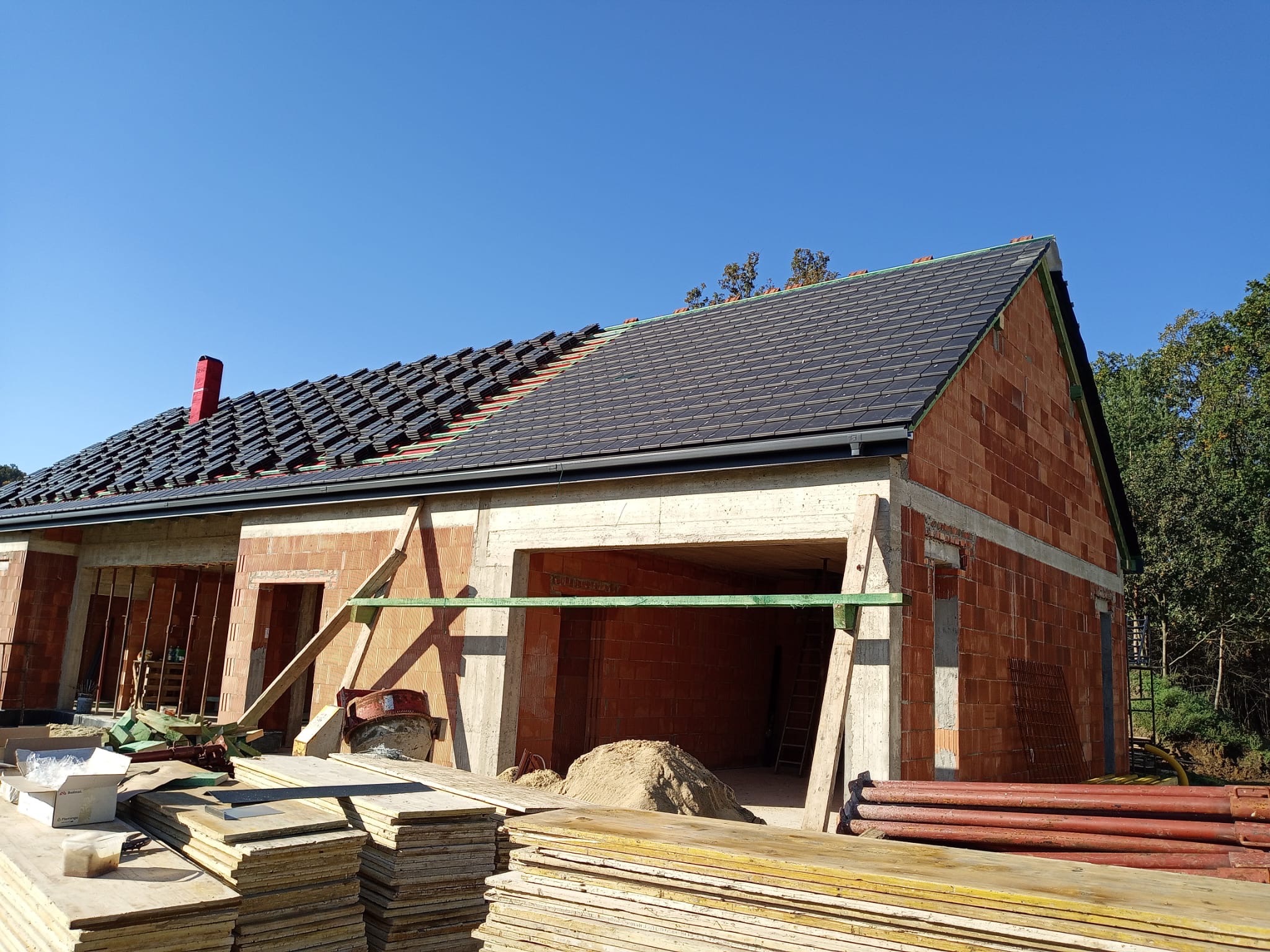 Niedokończony dom z czerwonej cegły z częściowo ułożonym dachem z czarnej dachówki i czerwonym kominem. Widoczne materiały budowlane i drewniane podpory.