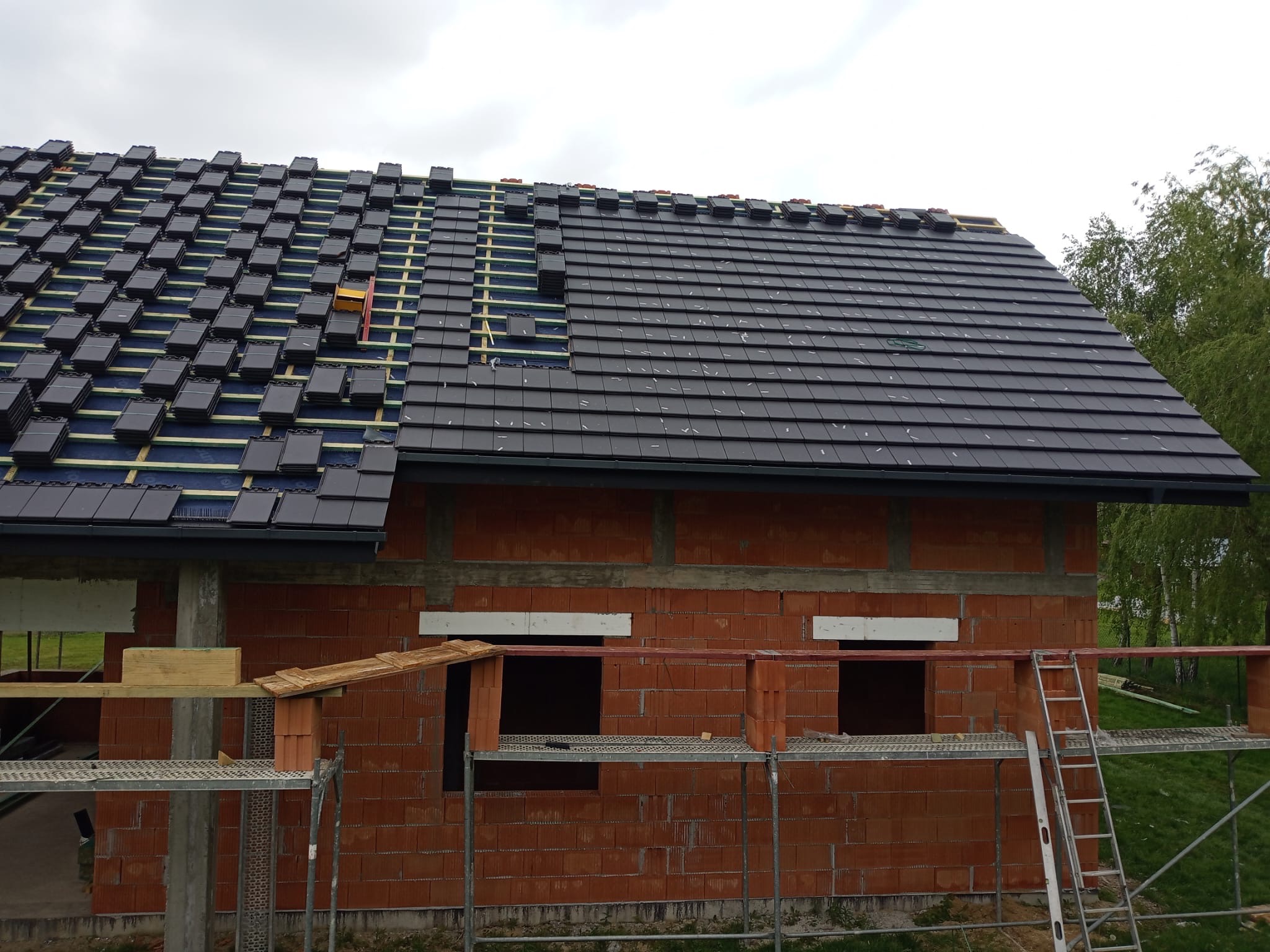 Budowa domu z czerwonej cegły: częściowo pokryty dach czarną dachówką, rusztowania i drabina przy ścianie. Widok na zielone drzewa w tle.