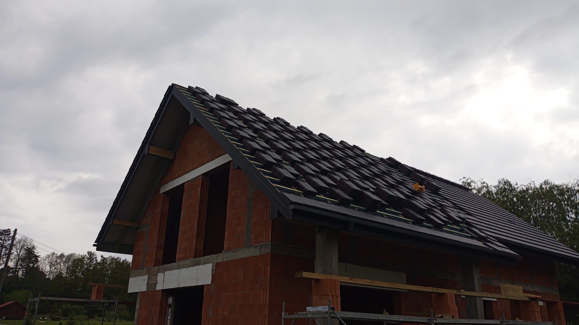 Wznoszenie domu z czerwonej cegły: widok na częściowo pokryty dach z ciemnej dachówki, okna z białymi nadprożami, pochmurne niebo w tle.