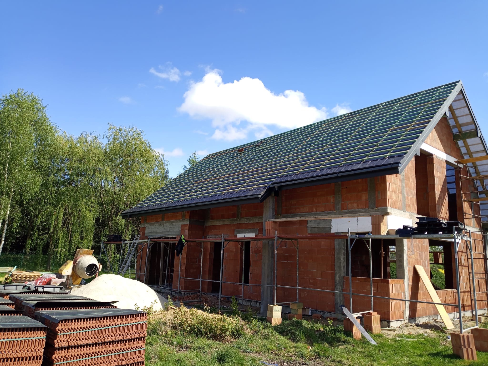 Murowany dom w trakcie budowy z częściowo położonym dachem, widoczne rusztowania, mieszarka i paleta z dachówkami na pierwszym planie.