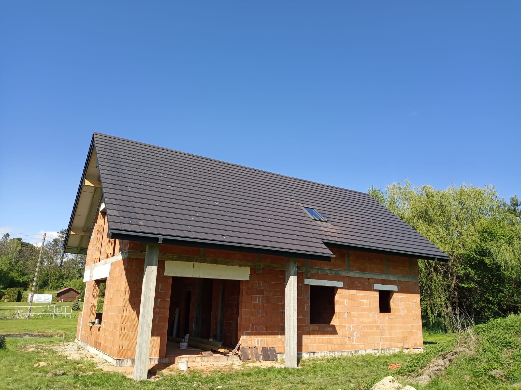 Niedokończony dom z czerwonej cegły z ciemnym dachem i oknem dachowym, otoczony zielenią i błękitnym niebem w słoneczny dzień. Widoczne betonowe filary.