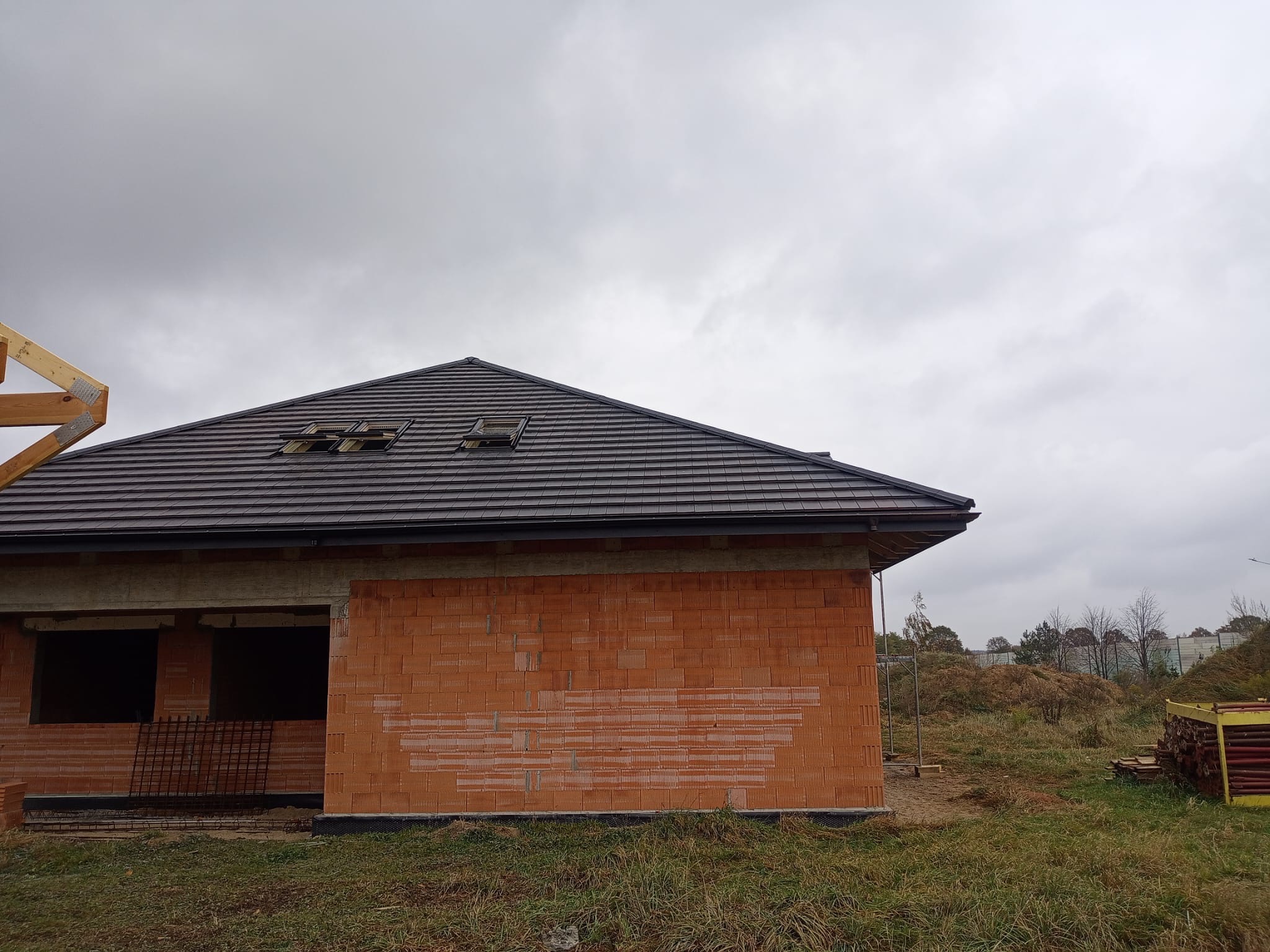 Niedokończony dom z czerwonej cegły pod ciemnym dachem z oknami dachowymi, widok z zewnątrz w pochmurny dzień. Częściowo widoczna konstrukcja drewniana.