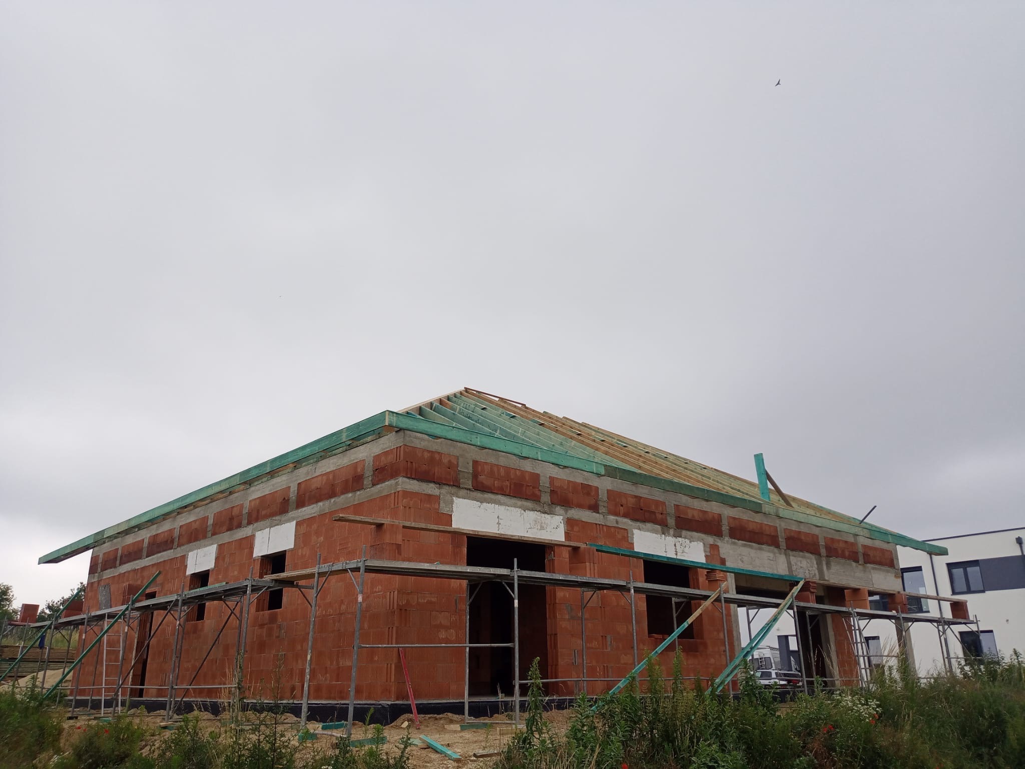 Budowa domu jednorodzinnego z czerwonej cegły, widoczne rusztowania i elementy konstrukcji dachu. Pochmurne niebo w tle, budynek w stanie surowym otwartym.