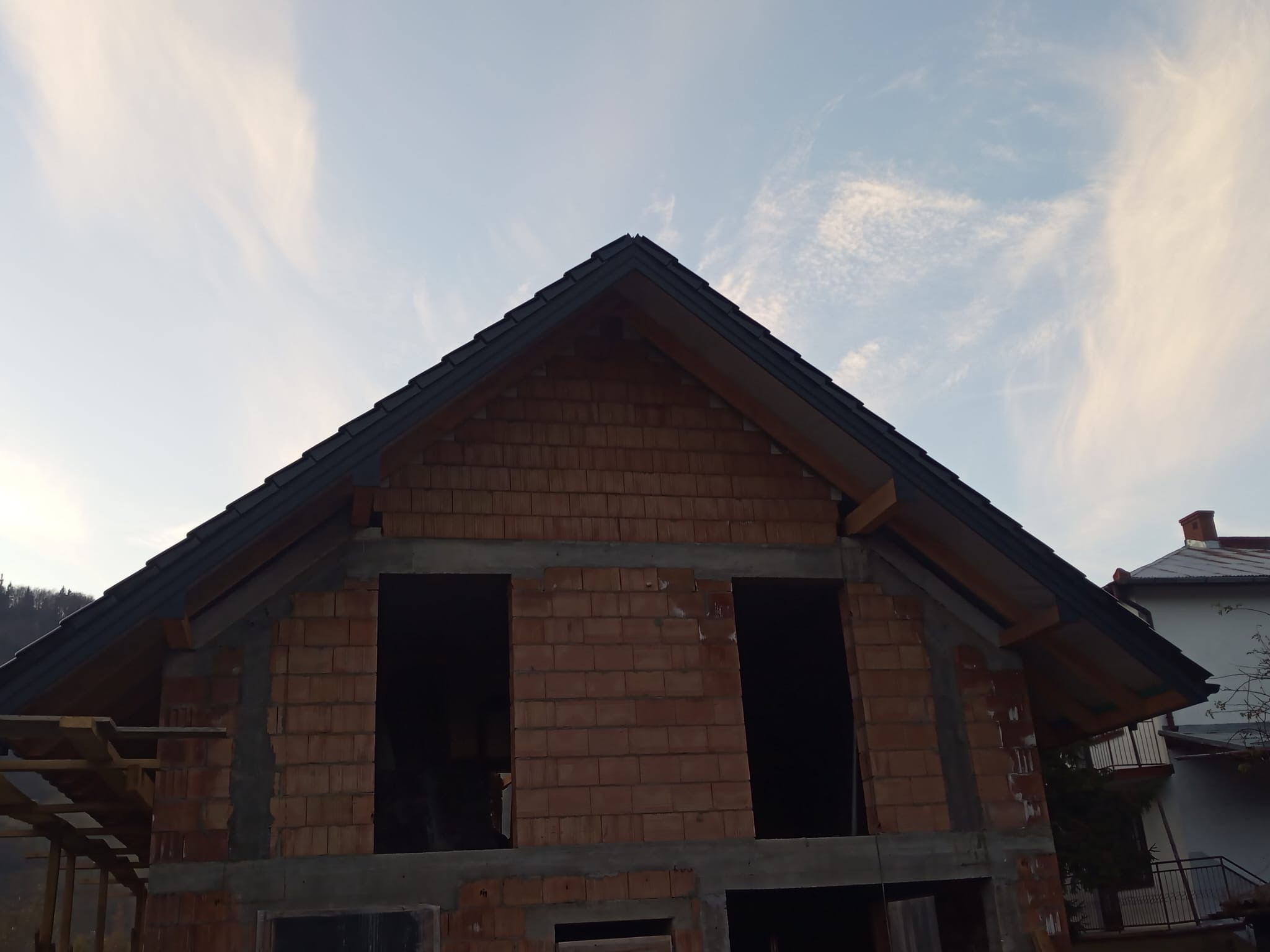 Surowy stan domu z cegły z widocznym dachem i otworami okiennymi na tle błękitnego nieba z chmurami. Widok z dołu na budynek w trakcie budowy.