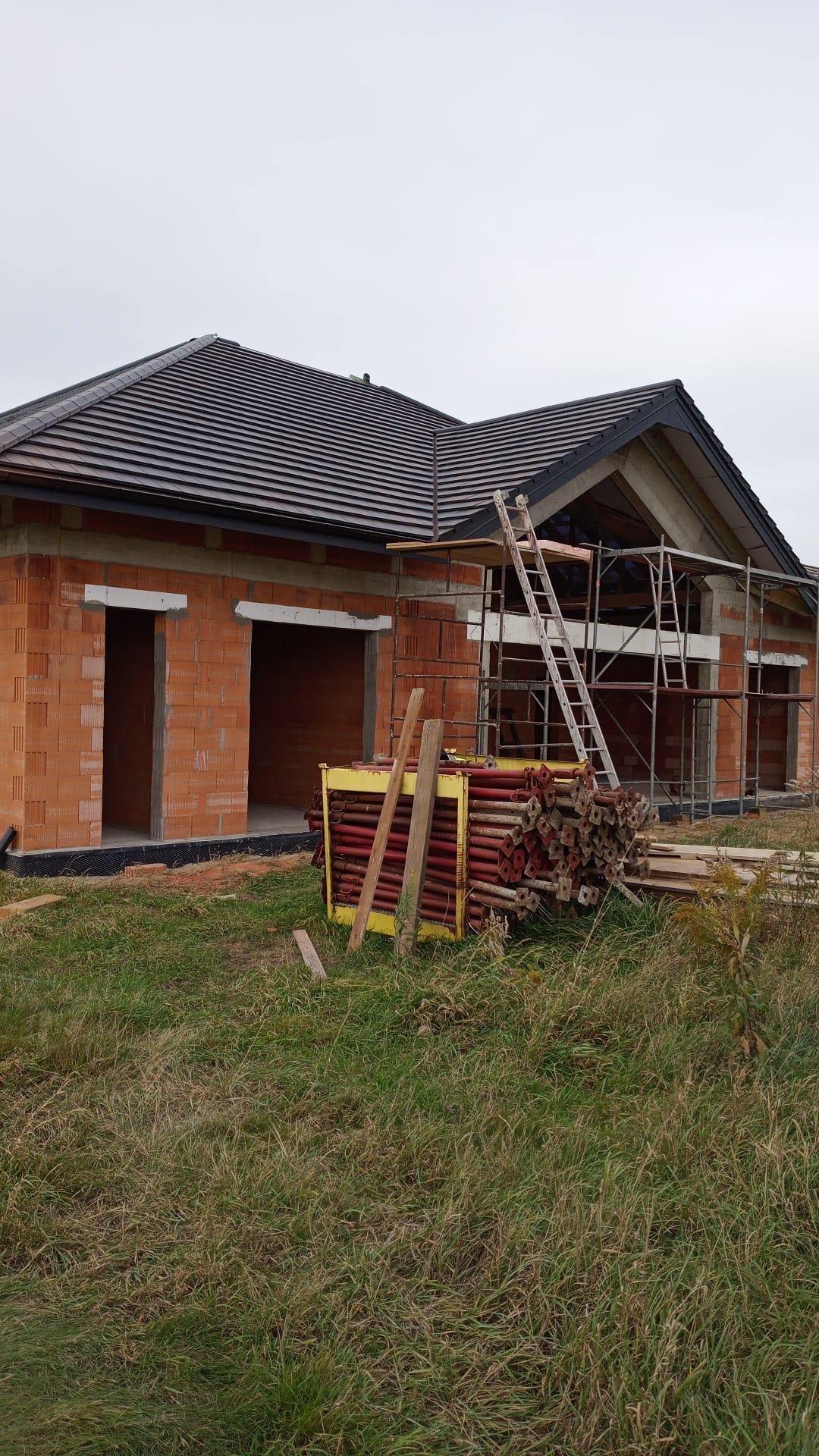 Budowa domu jednorodzinnego w trakcie: ściany z czerwonej cegły, dach z ciemnej dachówki, rusztowania. Na pierwszym planie skrzynia z podporami.