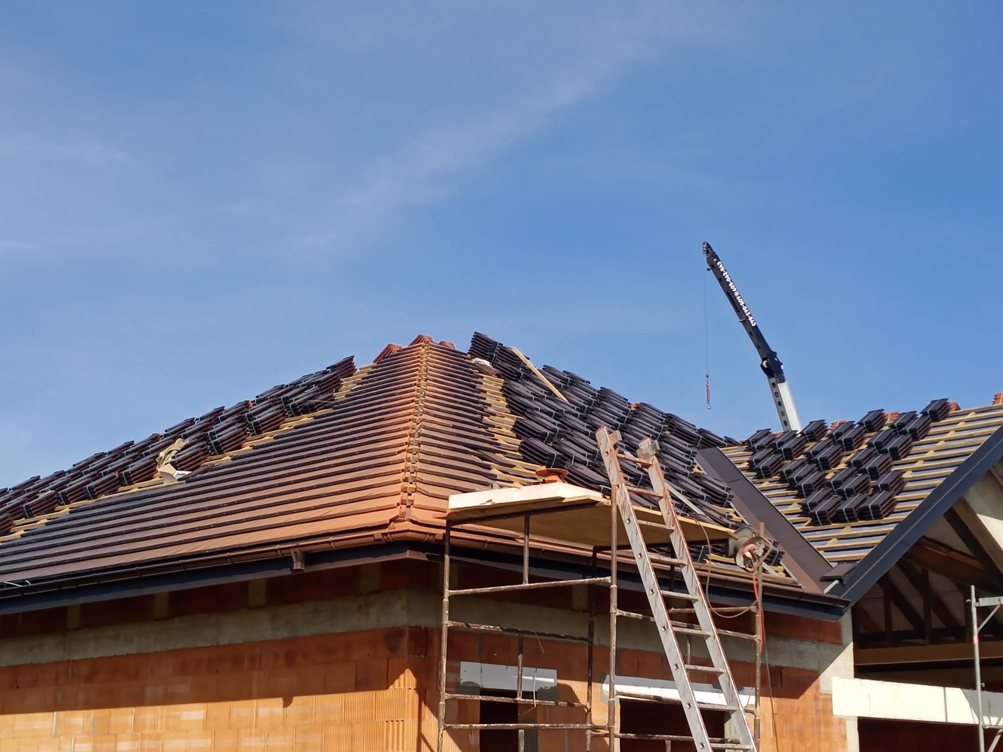 Układanie dachówki ceramicznej na dachu domu jednorodzinnego, widoczne rusztowanie i dźwig w tle, etap prac dekarskich w słoneczny dzień.