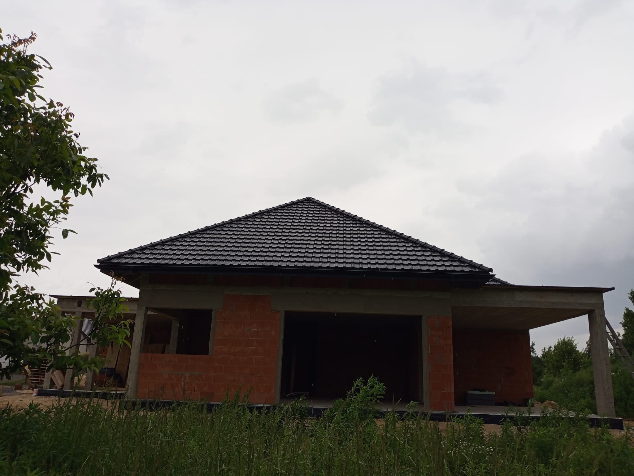 Surowy, murowany dom jednorodzinny z ciemną dachówką, w trakcie budowy, z widocznymi elementami konstrukcyjnymi i zielenią w tle, pochmurne niebo.