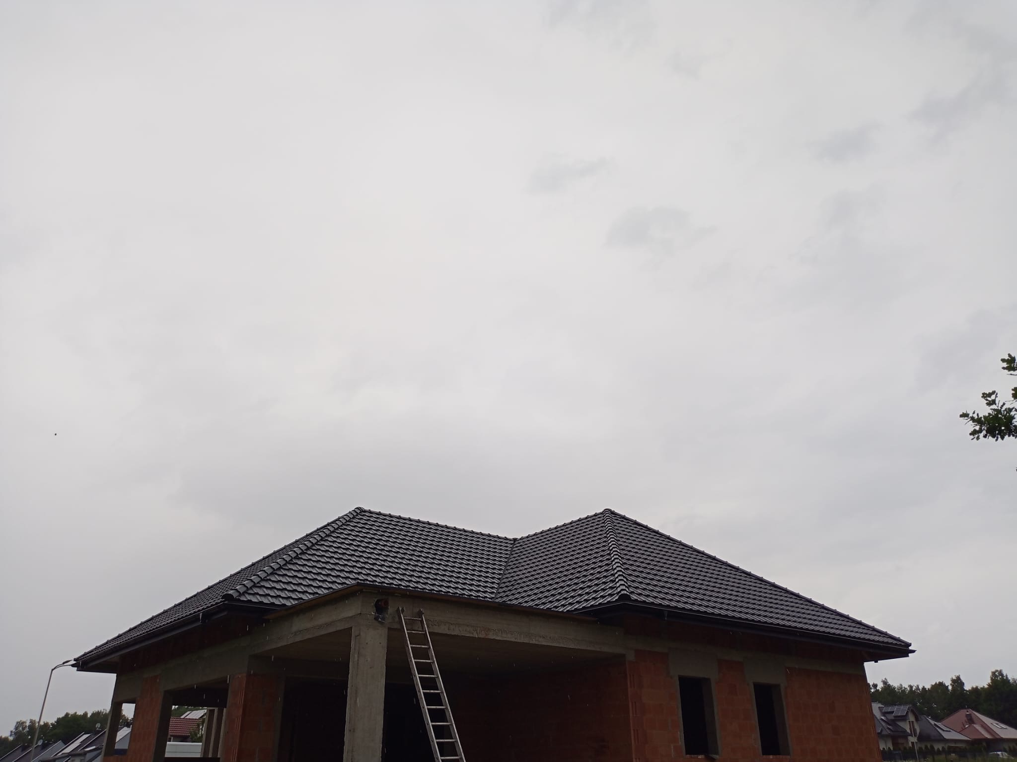 Dom w trakcie budowy z ciemną dachówką, widoczna drabina oparta o betonowy filar. Czerwona cegła na ścianach, pochmurne niebo w tle.