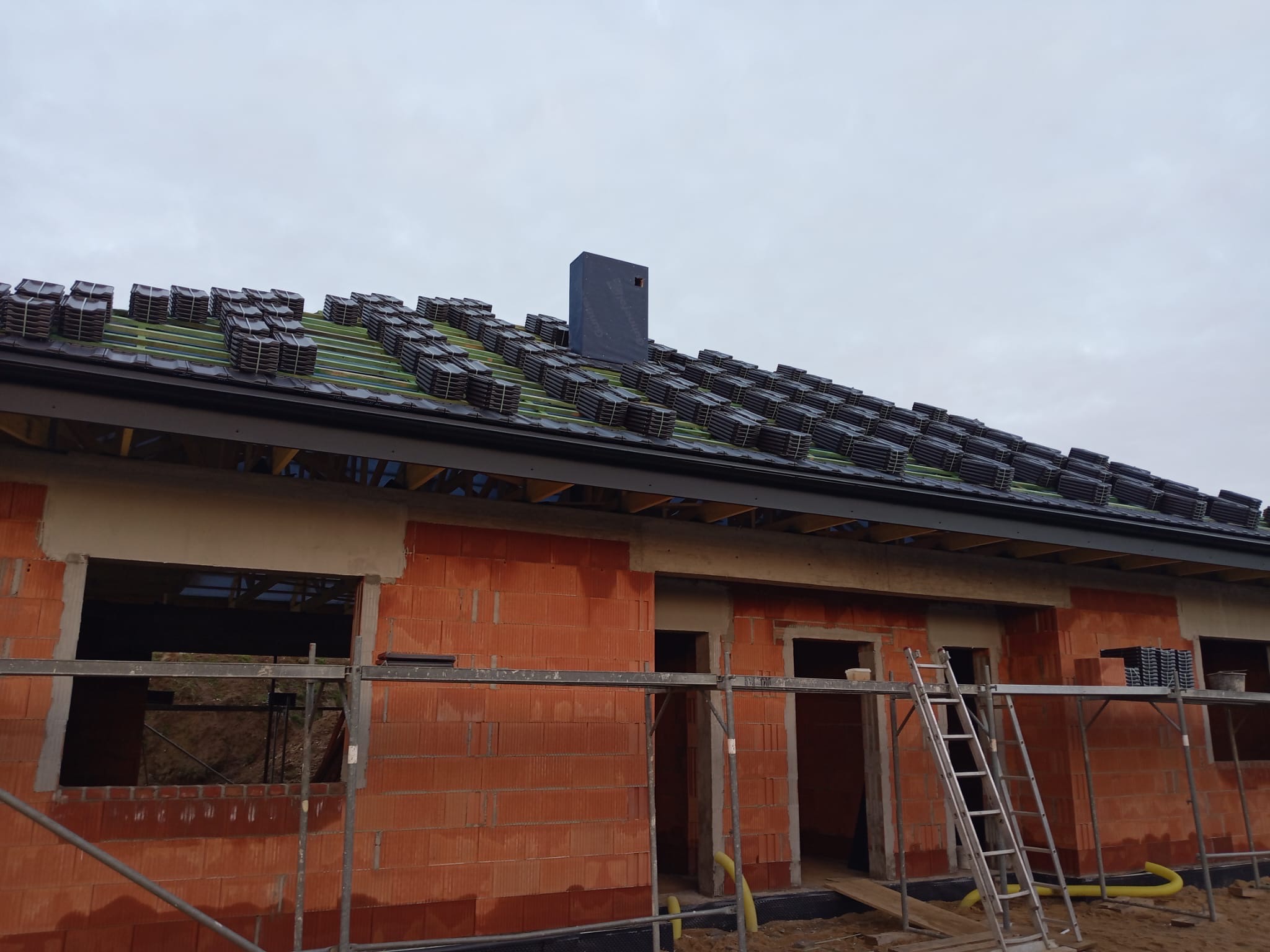 Niedokończony dom z czerwonej cegły z ułożonymi dachówkami na dachu. Widoczne rusztowania i drabina. Etap budowy w pochmurny dzień.