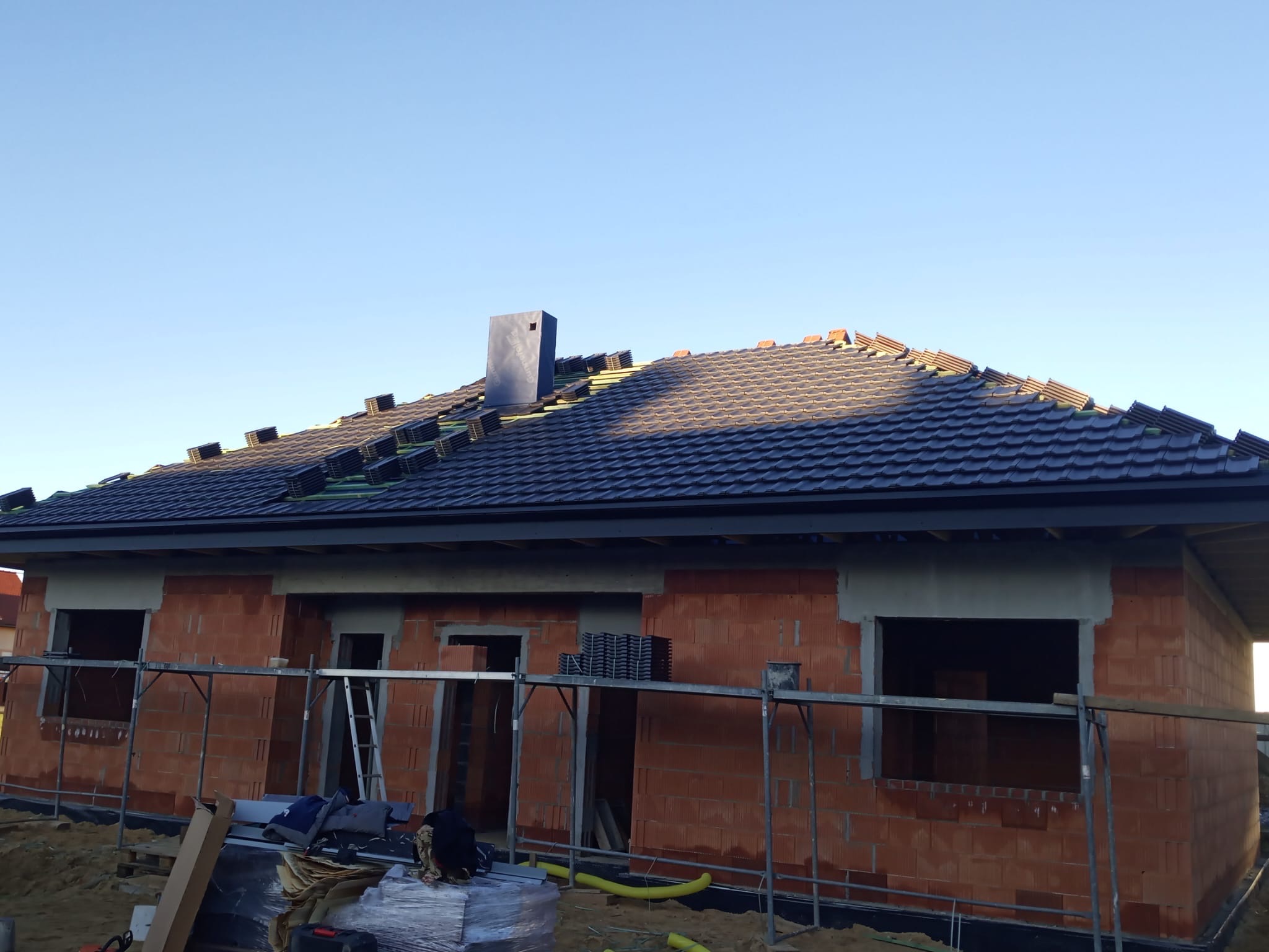 Budowa domu jednorodzinnego w trakcie realizacji: murowane ściany z pustaków, częściowo pokryty dach z czarnej dachówki, rusztowania. Widok z dołu na tle błękitnego nieba.