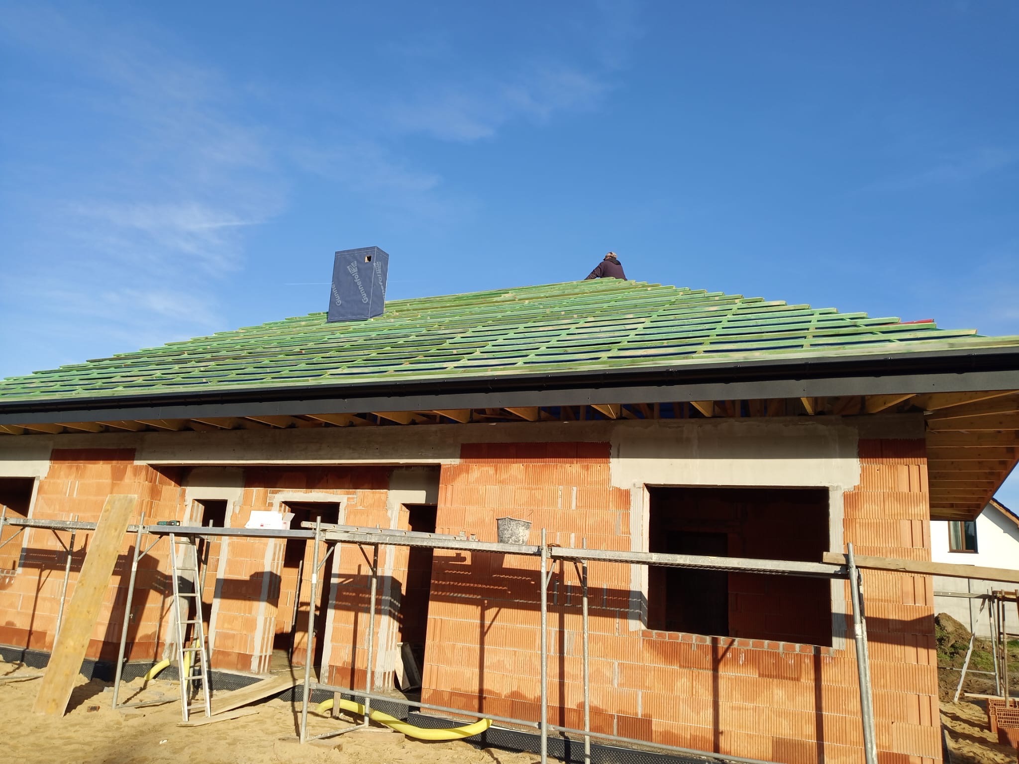 Budowa domu z czerwonej cegły: widoczne rusztowania, okna, zielony dach z kominem. Na dachu pracuje osoba. Błękitne niebo w tle.