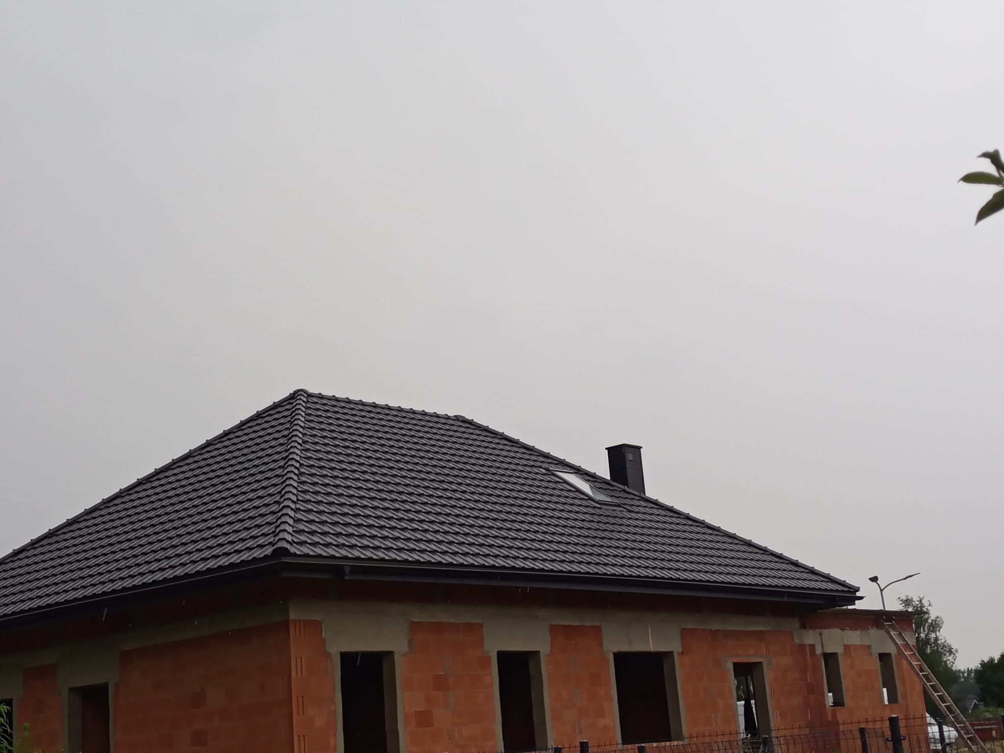 Surowy stan domu z cegły z czarnym dachem, widoczne otwory okienne i komin. Na zewnątrz drabina oparta o ścianę. Pochmurne niebo w tle.