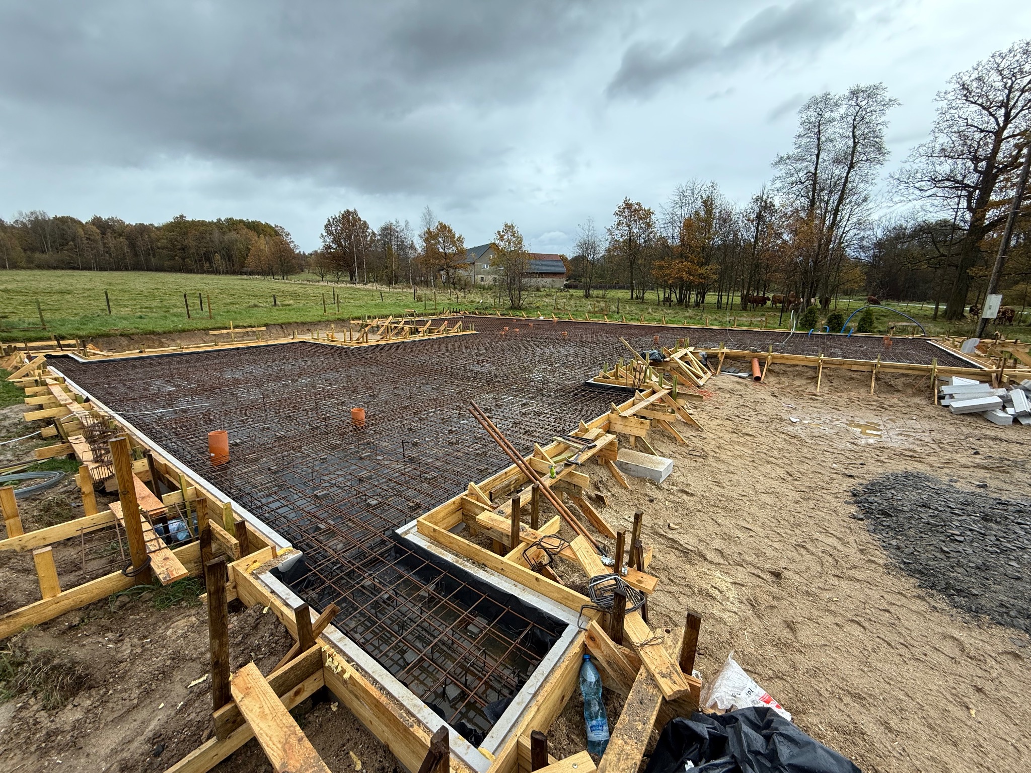 Zbrojenie fundamentów domu jednorodzinnego z drewnianym szalunkiem, widok z góry na rozległy teren budowy w pochmurny dzień. W tle zabudowania gospodarcze i drzewa.