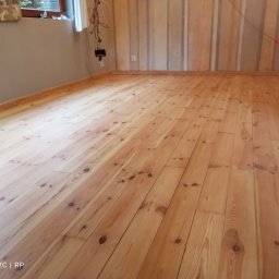 Renoparkiet - Jasna, drewniana podłoga z widocznymi słojami, świeżo po renowacji. Ściany w odcieniach szarości i beżu. Widoczne gniazdka elektryczne i kable.