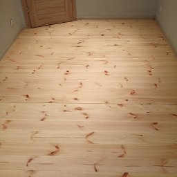 Renoparkiet - Jasna, drewniana podłoga z widocznymi słojami i sękami, ułożona w regularne deski. W tle fragment ściany i drzwi z jasnego drewna.