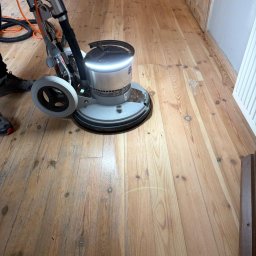 Renoparkiet - Cyklinowanie drewnianej podłogi maszyną z oświetleniem LED. Widoczny fragment podłogi po obróbce, jaśniejszy od reszty. Fragment butów operatora w lewym dolnym rogu.