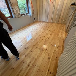 Renoparkiet - Błyszcząca, świeżo polakierowana podłoga z jasnego drewna w pokoju z oknami i tapetą w pionowe pasy. Widoczny fragment osoby w ochraniaczach na butach.