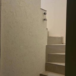 DAM BUD - Wykończona klatka schodowa z dekoracyjną ścianą o fakturze trawertynu i schodami wyłożonymi płytkami w odcieniach beżu. Minimalistyczny design, neutralna kolorystyka.