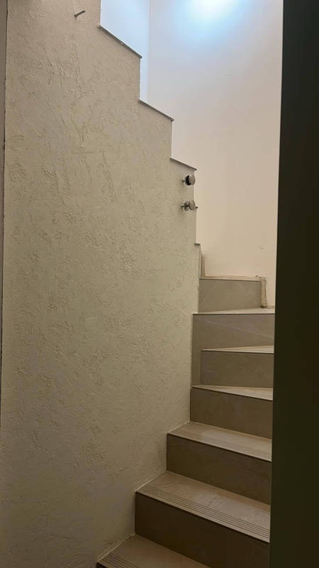 Wykończona klatka schodowa z dekoracyjną ścianą o fakturze trawertynu i schodami wyłożonymi płytkami w odcieniach beżu. Minimalistyczny design, neutralna kolorystyka.