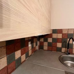 Eryk Kupis - Jasne, drewniane szafki kuchenne nad blatem z szarego kamienia i mozaiką z kolorowych płytek. Widoczny kran i gniazdko elektryczne.