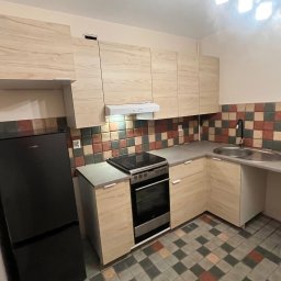 Eryk Kupis - Jasna, drewnopodobna zabudowa kuchenna z czarną lodówką i płytą grzewczą na tle mozaiki ściennej i podłogowej. Ujęcie szerokokątne.