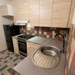 Eryk Kupis - Jasna kuchnia z drewnianymi szafkami, czarną lodówką i mozaiką nad blatem. Ujęcie z perspektywy zlewu, ukazujące detale wykończenia i aranżacji.