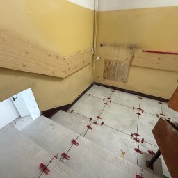 Scarbud - Oskar Kolaska - Kładzenie jasnych płytek na schodach z użyciem czerwonych klipsów poziomujących. Żółte ściany i drewniane poręcze. Widok z góry na etap prac wykończeniowych.