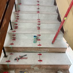 Scarbud - Oskar Kolaska - Układanie płytek na schodach z użyciem systemu poziomowania. Widoczne narzędzia, poziomica i puszka napoju. Jasne płytki schodowe w trakcie montażu.