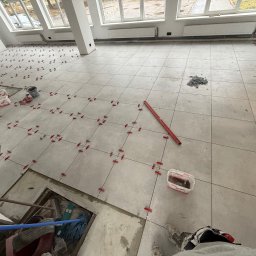 Scarbud - Oskar Kolaska - Układanie jasnych płytek podłogowych w dużym pomieszczeniu, z użyciem czerwonych klipsów poziomujących. Widoczne narzędzia i fragmenty nieukończonej podłogi. Duże okna w tle.