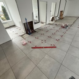 Scarbud - Oskar Kolaska - Układanie jasnych, kwadratowych płytek podłogowych w przestronnym wnętrzu. Widoczne poziomica, krzyżyki dystansowe i fragmenty niezakończonej podłogi.
