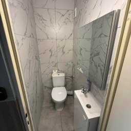 Scarbud - Oskar Kolaska - Wąska łazienka po remoncie: biała toaleta, umywalka z szafką i lustro. Ściany wyłożone płytkami imitującymi marmur. Widok zza futryny.