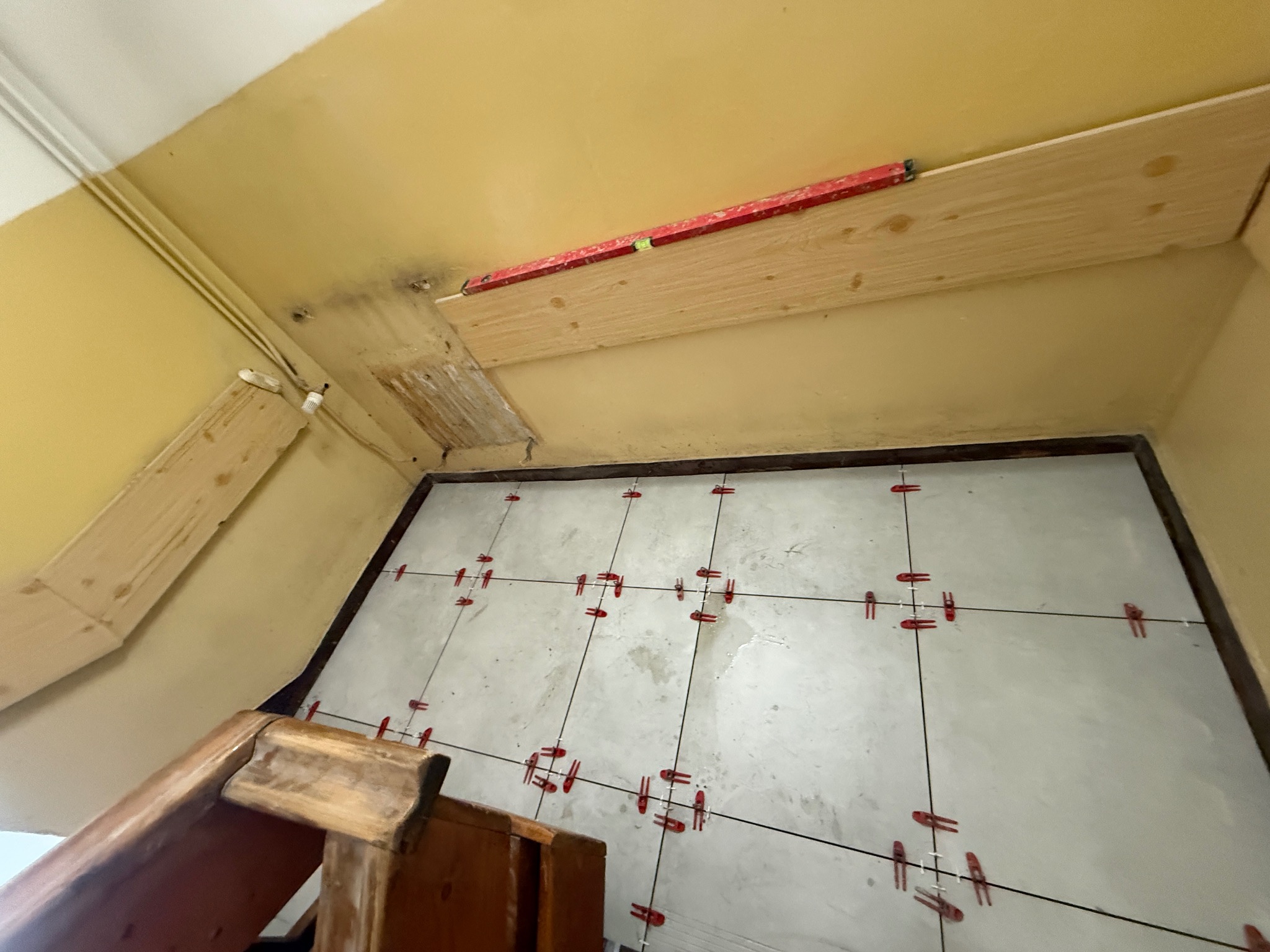 Przygotowanie do układania płytek: poziomowanie sufitu z użyciem poziomicy i listwy, widoczne krzyżyki dystansowe na podłodze. Ujęcie z dołu, fragment schodów.
