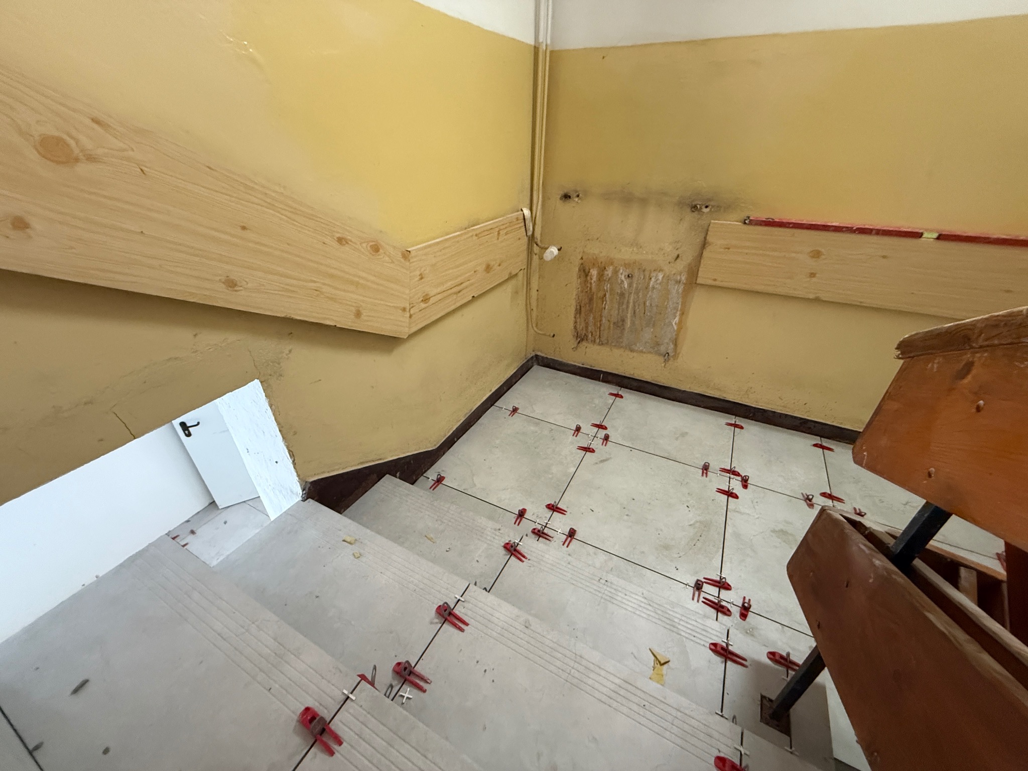 Kładzenie jasnych płytek na schodach z użyciem czerwonych klipsów poziomujących. Żółte ściany i drewniane poręcze. Widok z góry na etap prac wykończeniowych.