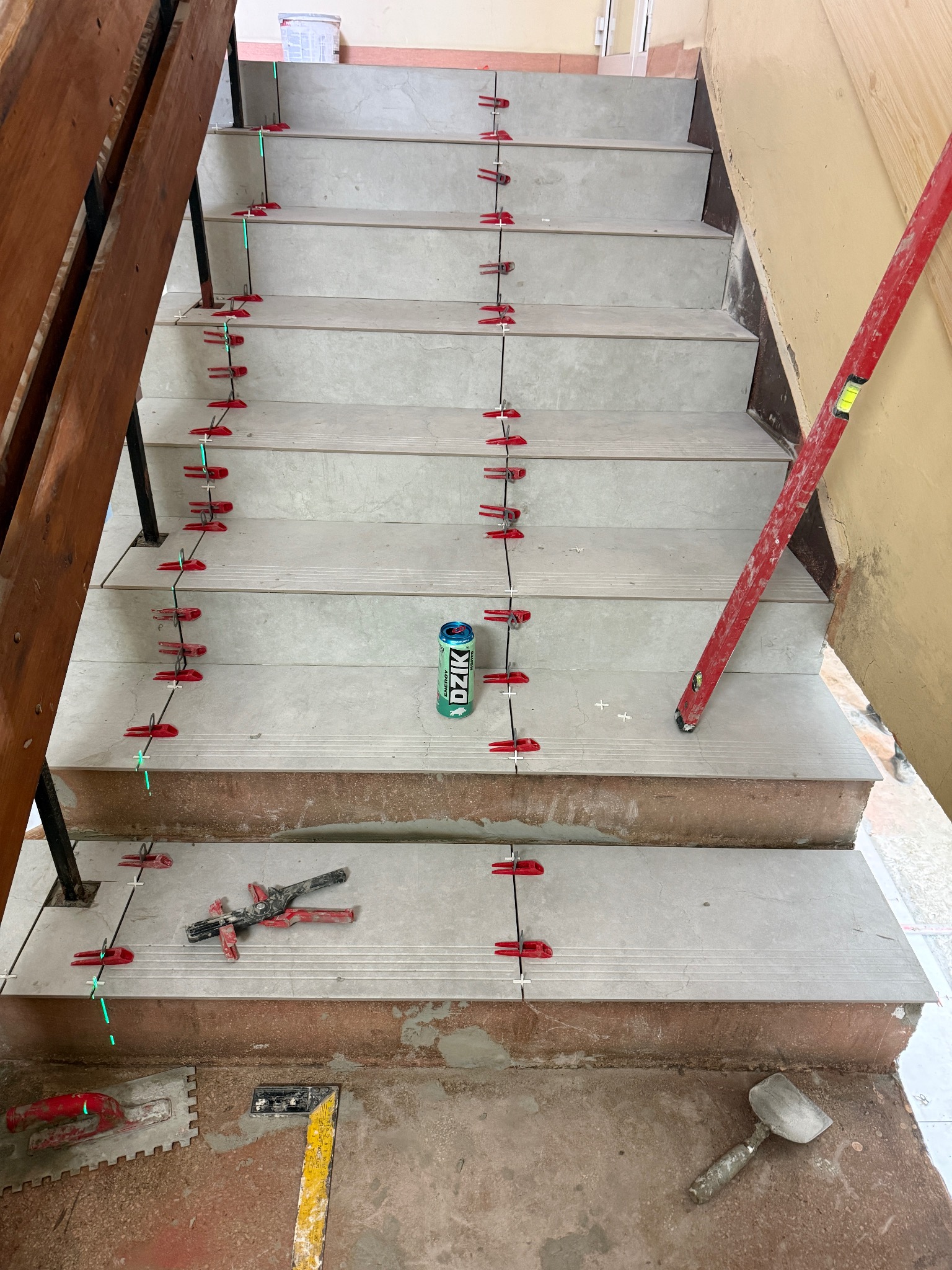 Układanie płytek na schodach z użyciem systemu poziomowania. Widoczne narzędzia, poziomica i puszka napoju. Jasne płytki schodowe w trakcie montażu.