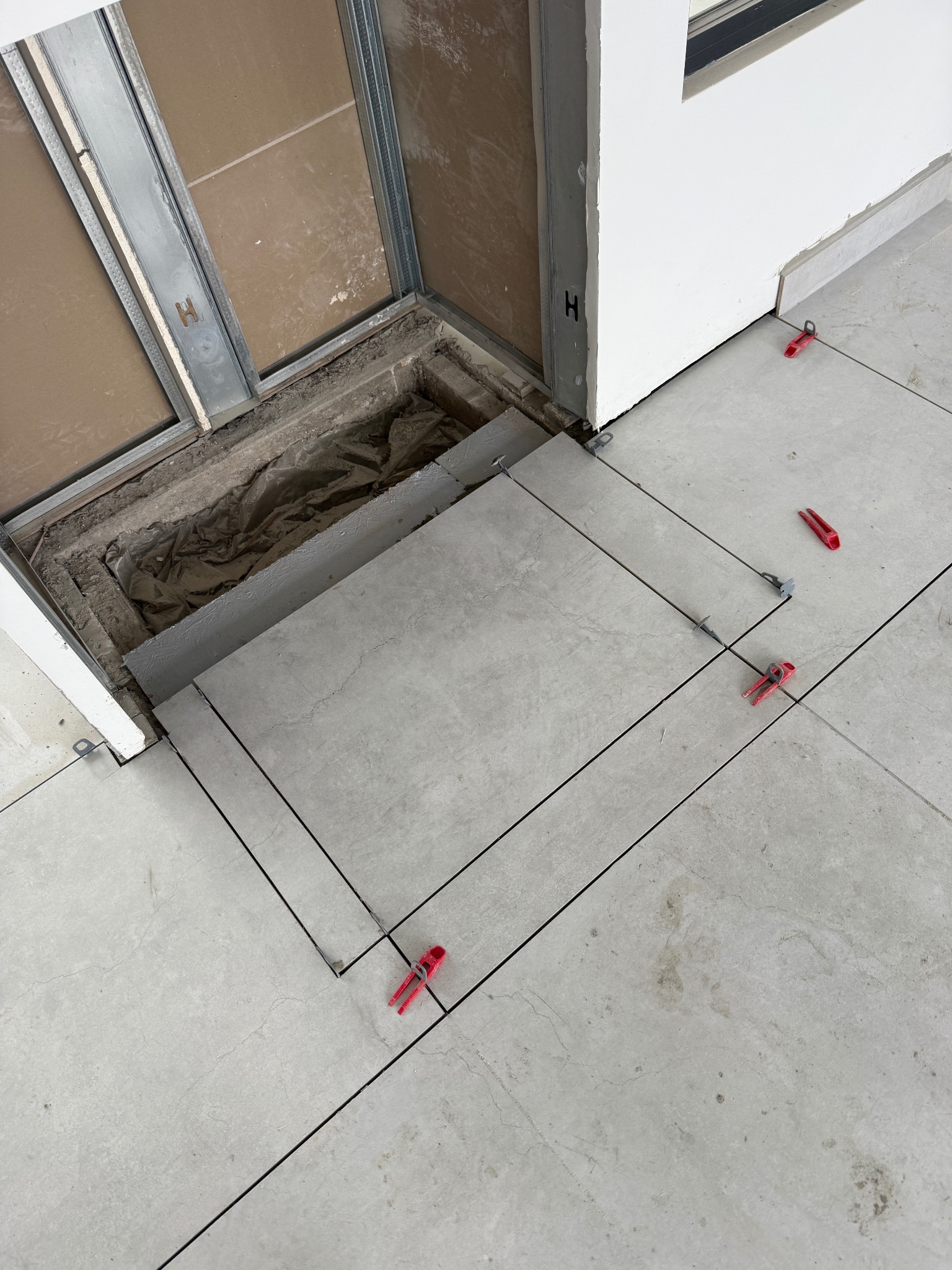 Układanie jasnoszarych płytek podłogowych w pomieszczeniu, widoczne czerwone klipsy poziomujące i fragment otworu w podłodze przy metalowej ramie drzwi.