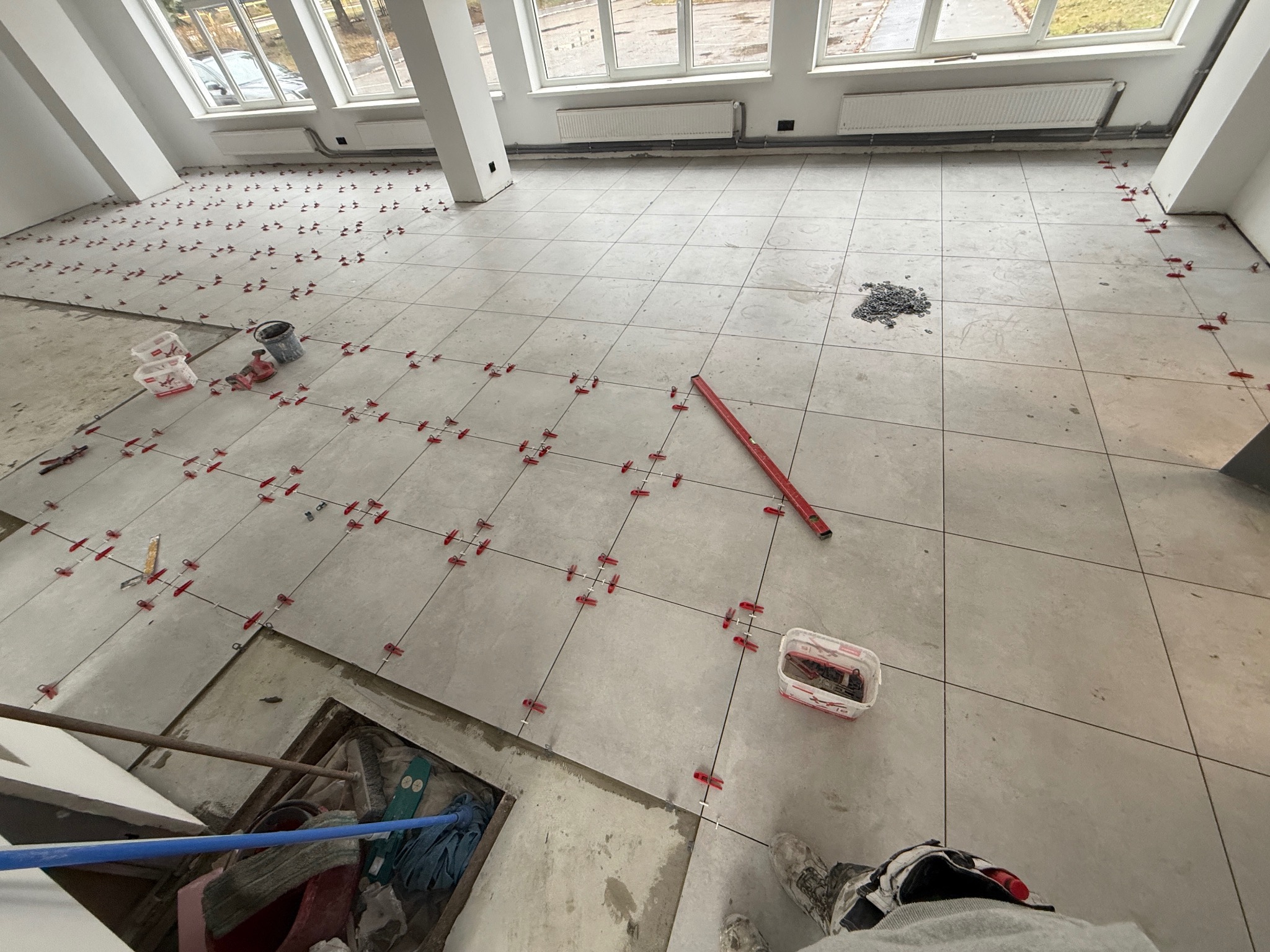 Układanie jasnych płytek podłogowych w dużym pomieszczeniu, z użyciem czerwonych klipsów poziomujących. Widoczne narzędzia i fragmenty nieukończonej podłogi. Duże okna w tle.