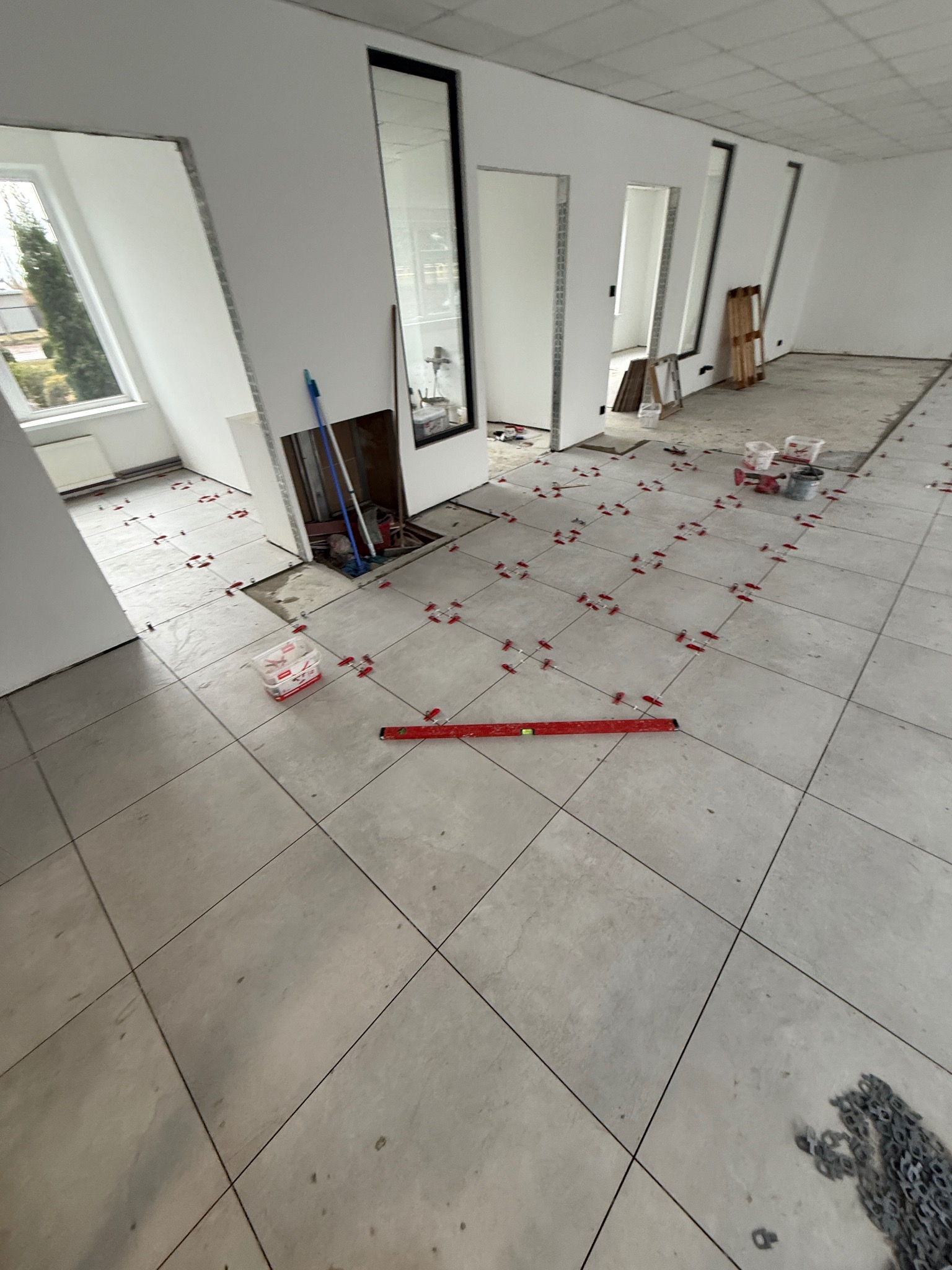 Układanie jasnych, kwadratowych płytek podłogowych w przestronnym wnętrzu. Widoczne poziomica, krzyżyki dystansowe i fragmenty niezakończonej podłogi.
