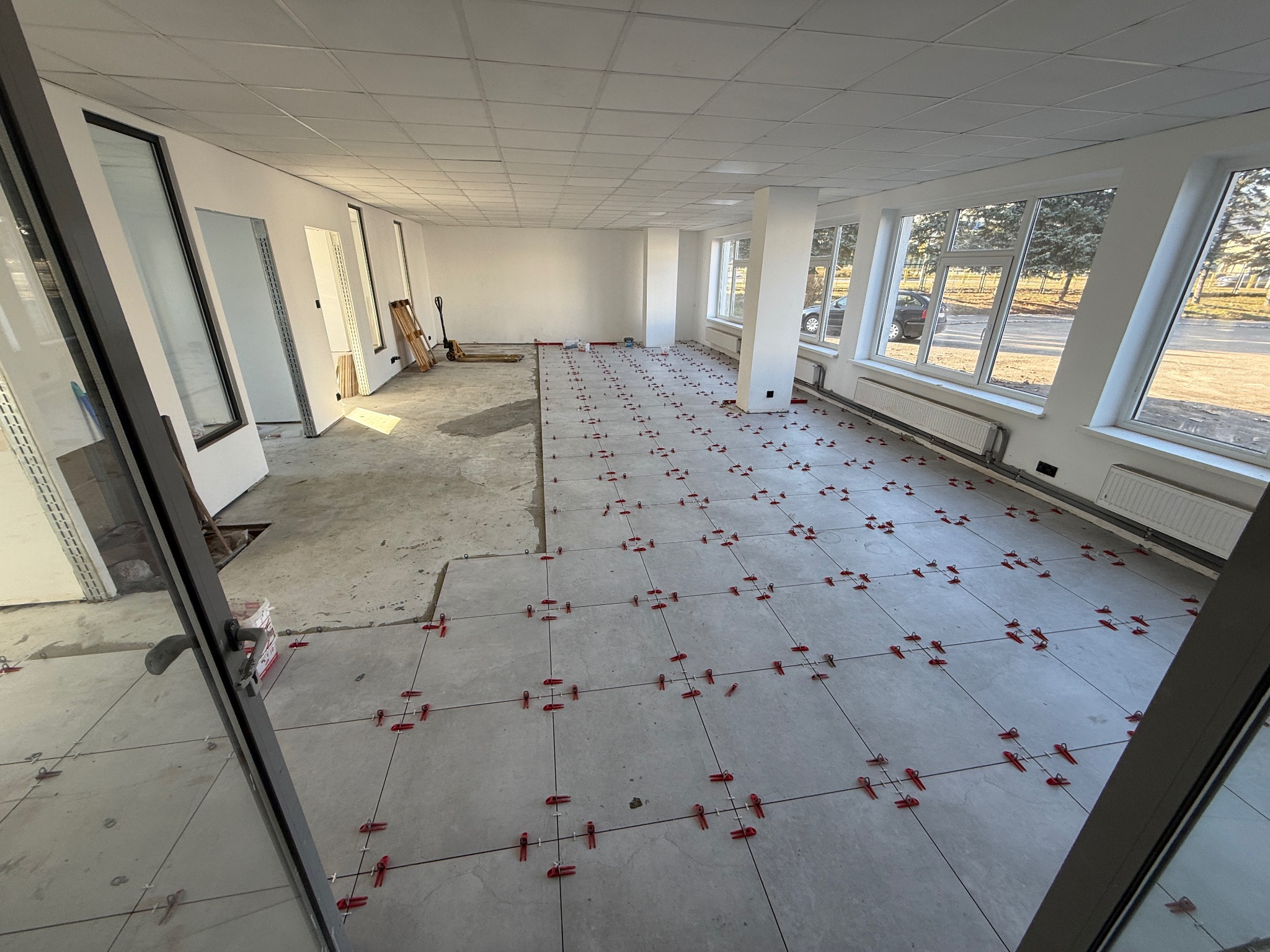 Wnętrze w trakcie układania jasnych, kwadratowych płytek podłogowych z czerwonymi krzyżykami dystansowymi. Część podłogi jeszcze nieukończona, widoczne narzędzia.