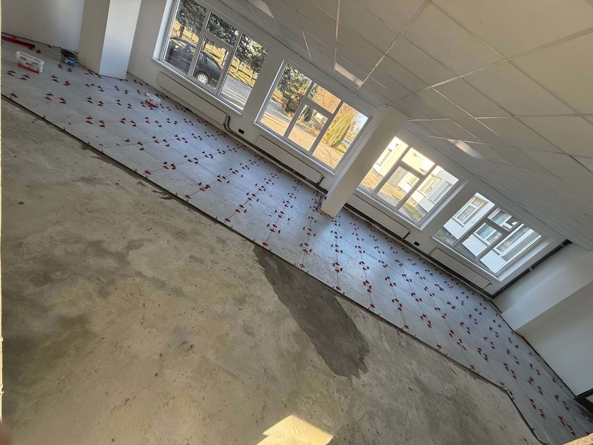 Układanie płytek podłogowych w jasnym pomieszczeniu z oknami. Widoczne krzyżyki dystansowe i surowa wylewka. Ujęcie z góry, ukośne.