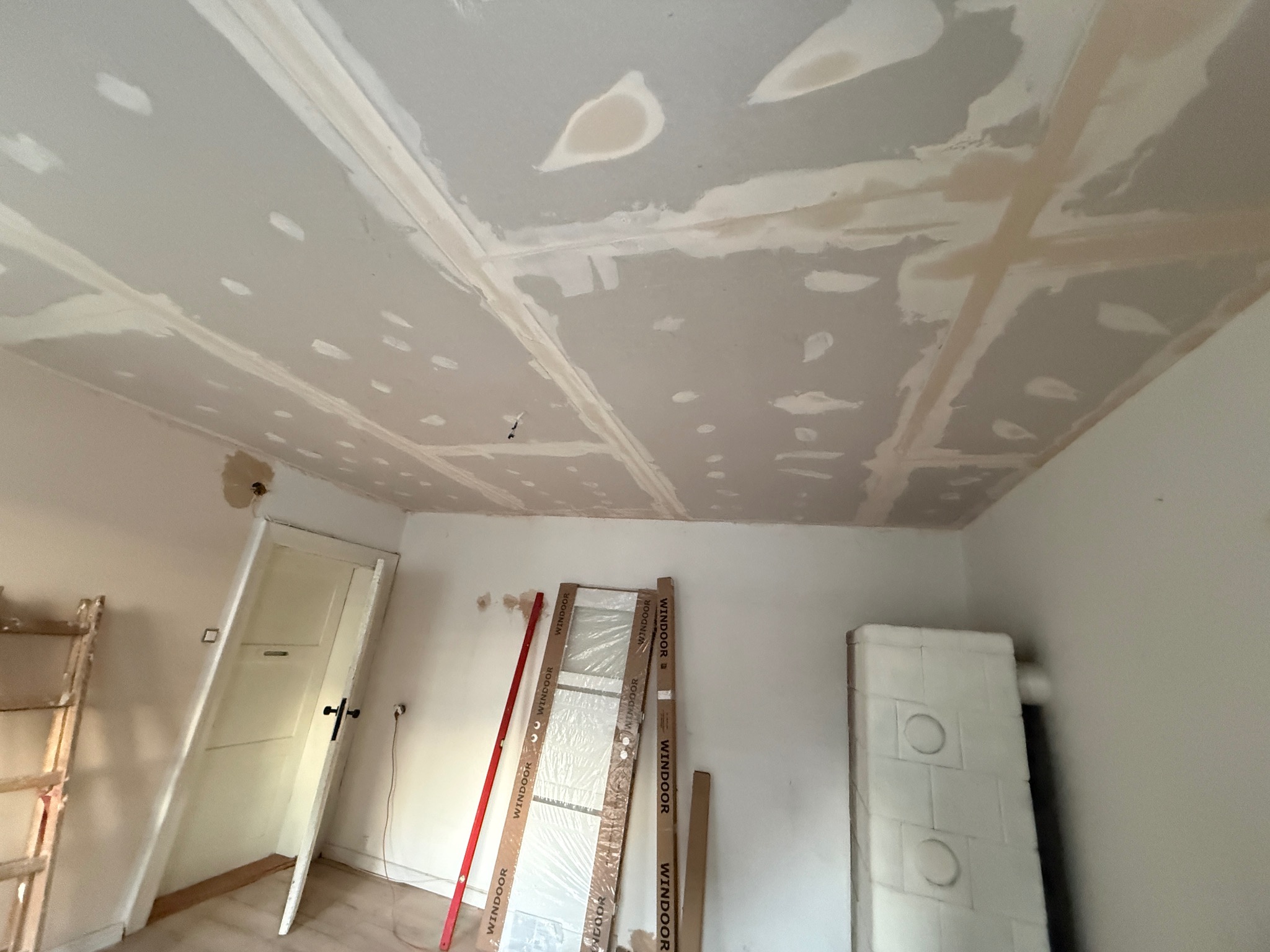 Sufit z płyt karton-gips, częściowo wyszpachlowany, z widocznymi łączeniami. W tle drzwi, grzejnik i drabina. Prace wykończeniowe w jasnym pomieszczeniu.