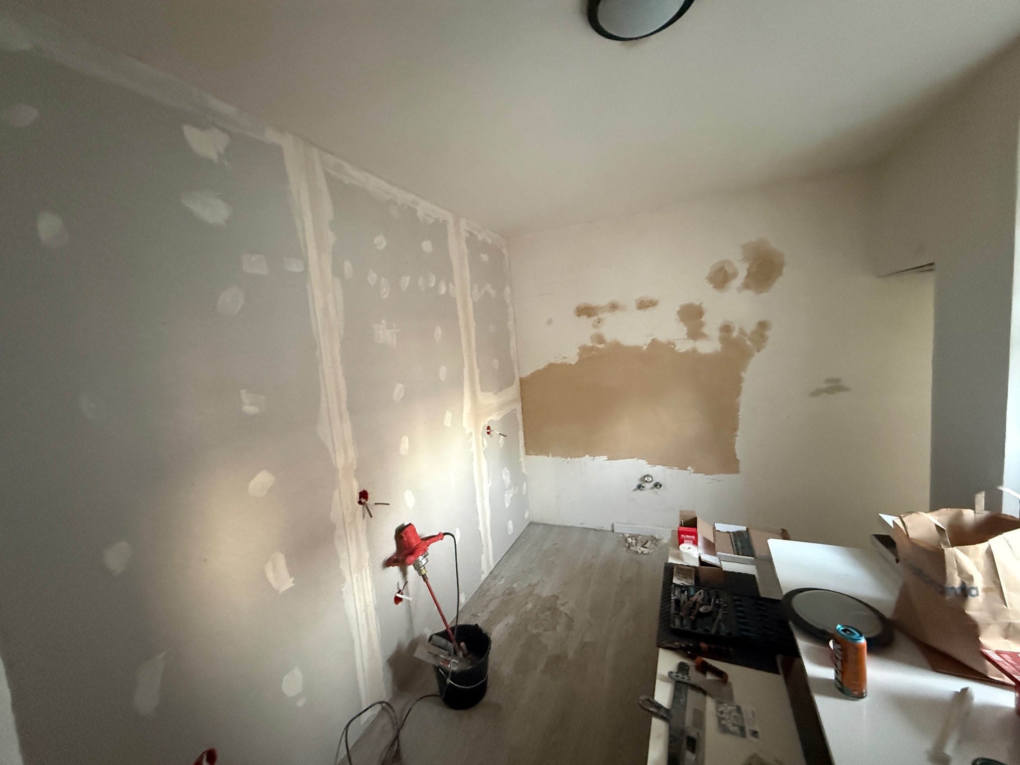 Ściana z płytami gipsowo-kartonowymi w trakcie szpachlowania, widoczne łączenia i punktowe wypełnienia. Na podłodze stoi mieszadło do gipsu. Częściowo przygotowana ściana do malowania.