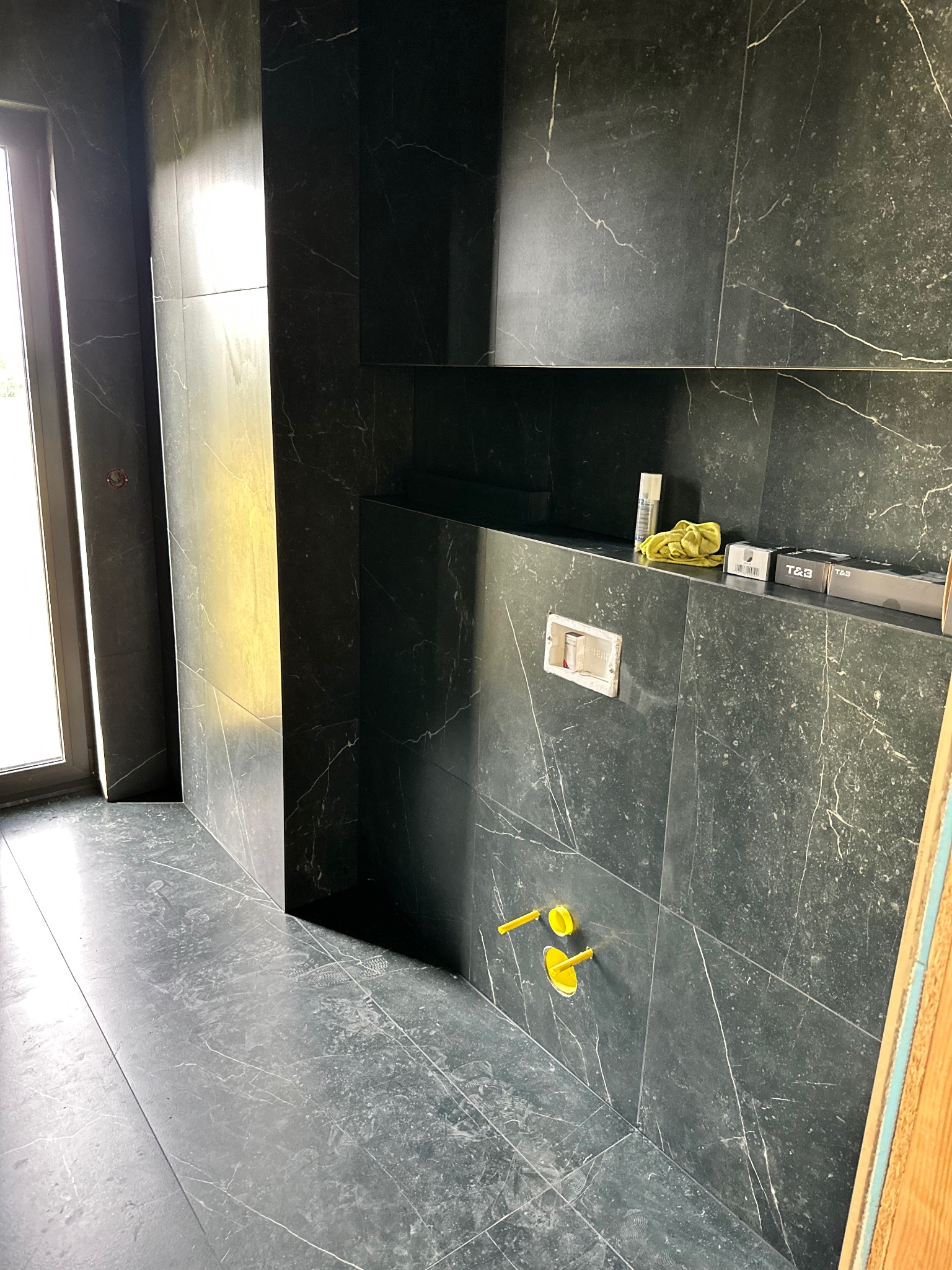 Wykończona łazienka z czarnymi płytkami imitującymi marmur, widoczne żółte elementy instalacji wodnej i półka z akcesoriami. Nowoczesny design, minimalistyczny styl.