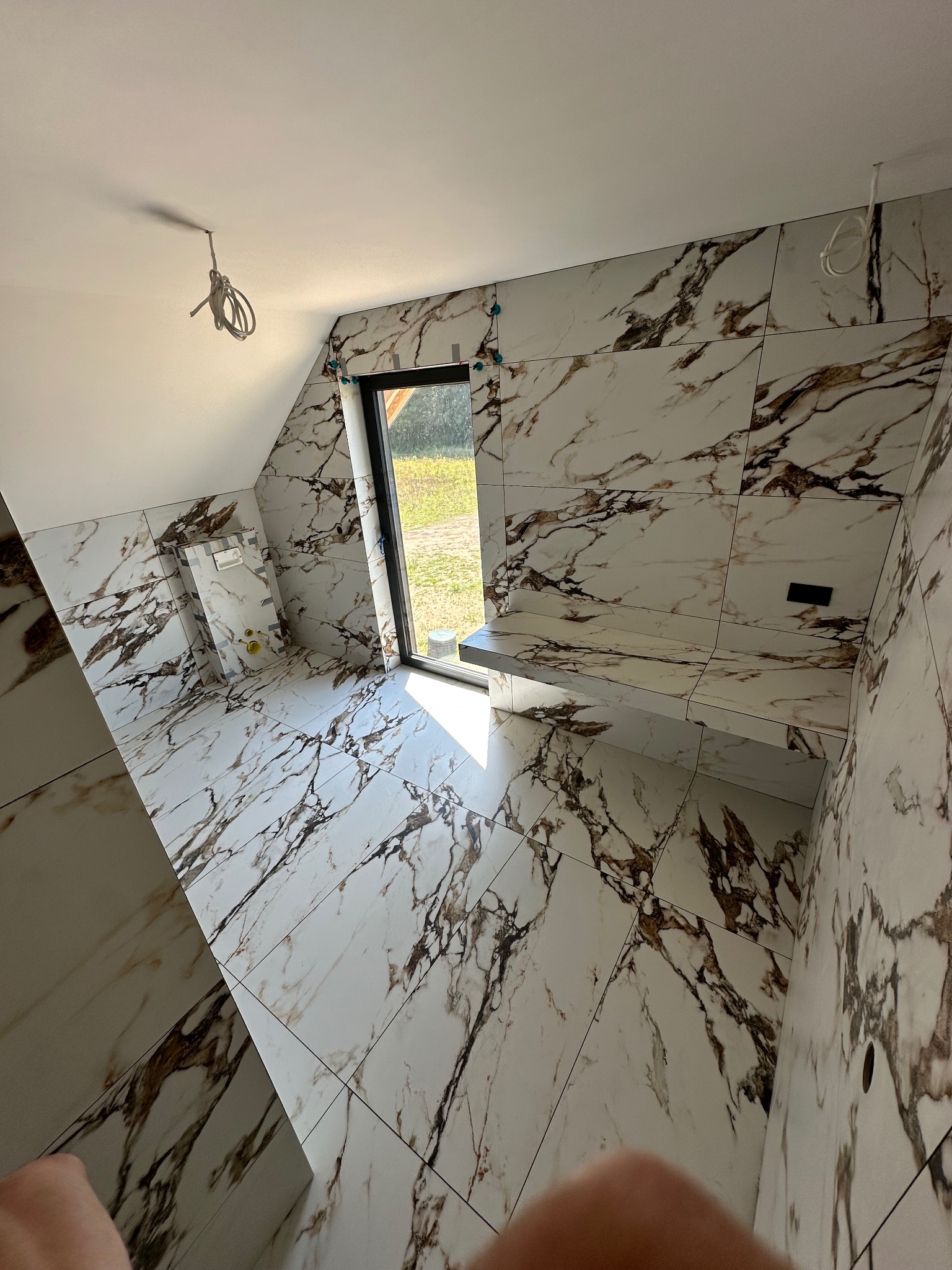 Ukończona łazienka na poddaszu z marmurowymi płytkami, widok z góry. Widoczne okno, zabudowa WC i fragment instalacji elektrycznej. Całość w jasnej tonacji.