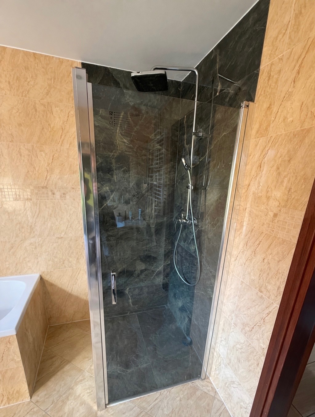 Nowoczesna kabina prysznicowa z ciemnego marmuru, chromowane wykończenia, beżowe płytki na ścianach i podłodze. Widok z boku, fragment wanny po lewej.
