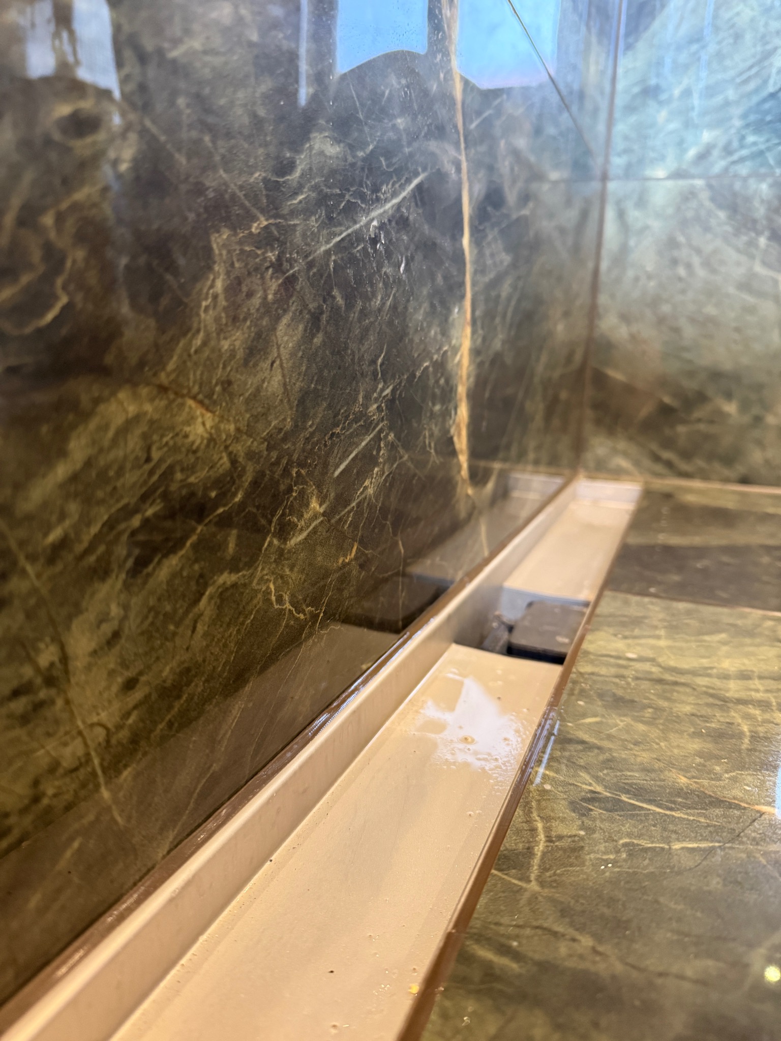 Ukończony odpływ liniowy w kabinie prysznicowej z marmurowymi płytkami w odcieniach zieleni i brązu. Widoczne detale wykończenia i połysk płytek.