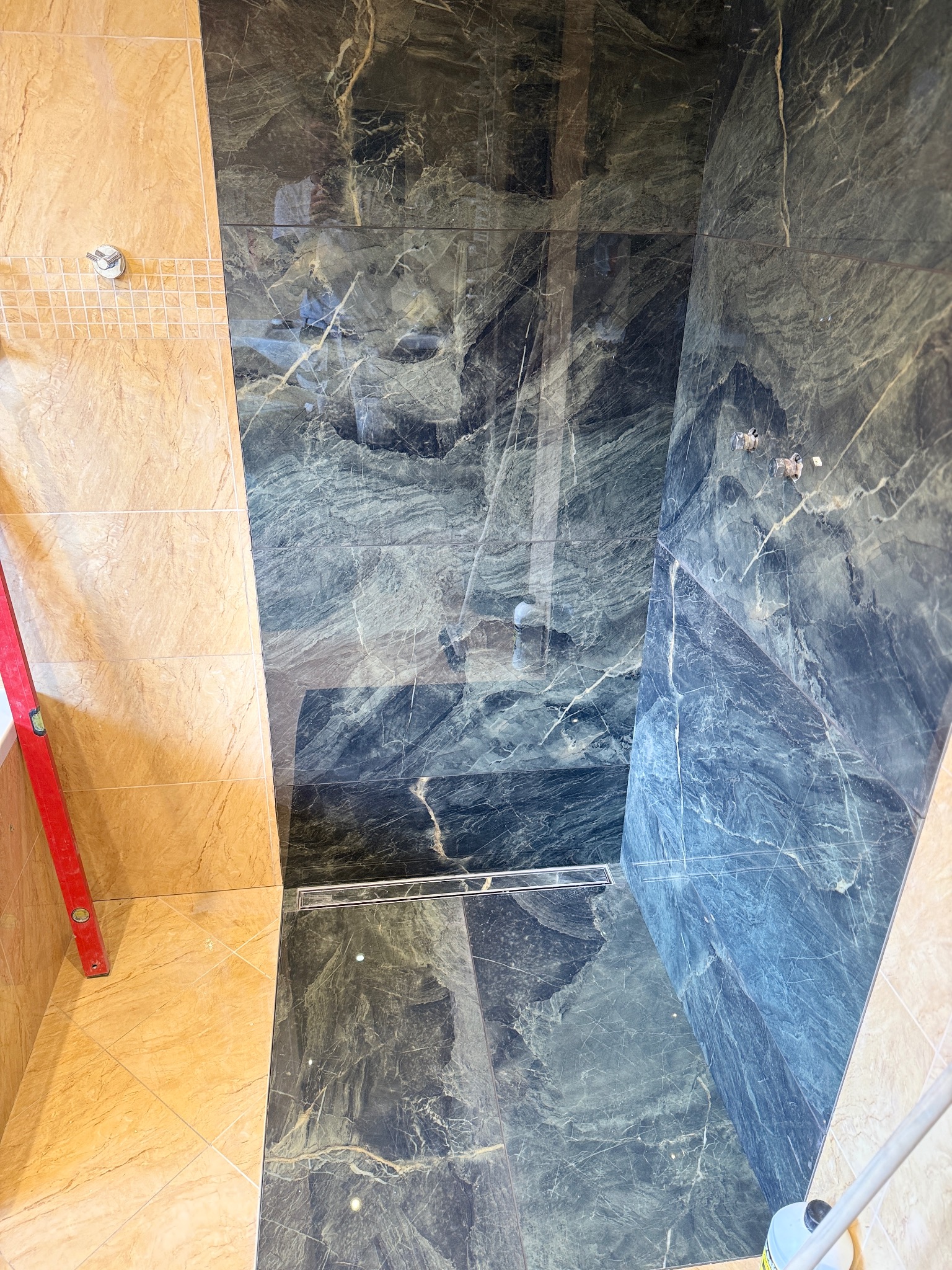 Wykończona kabina prysznicowa z ciemnymi płytkami imitującymi marmur i jasnymi płytkami po bokach. Widoczny odpływ liniowy i poziomica.