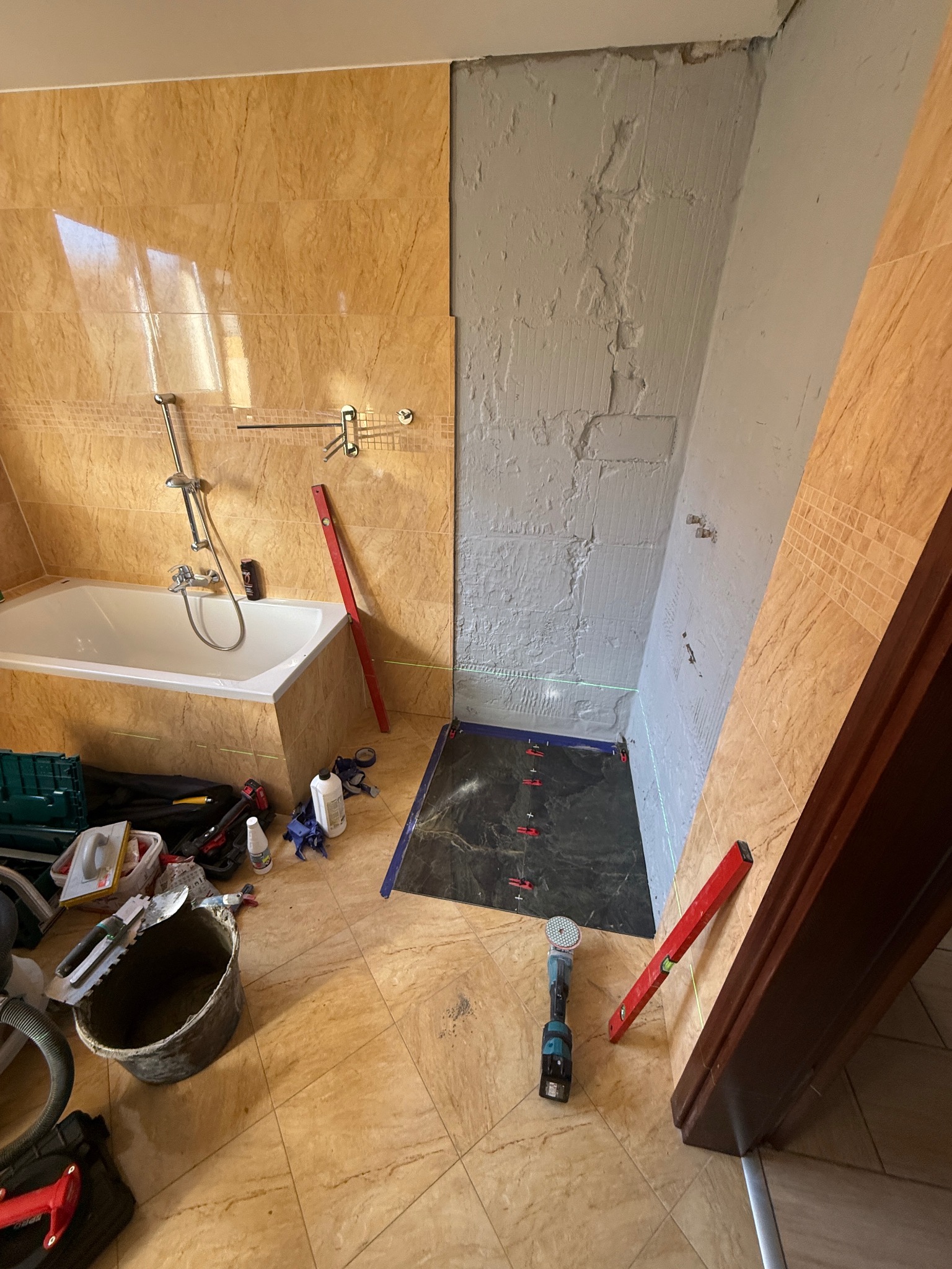 Łazienka w trakcie prac: częściowo wyłożone płytki na ścianach, przygotowane podłoże pod prysznic, narzędzia i poziomice widoczne na podłodze z płytek.