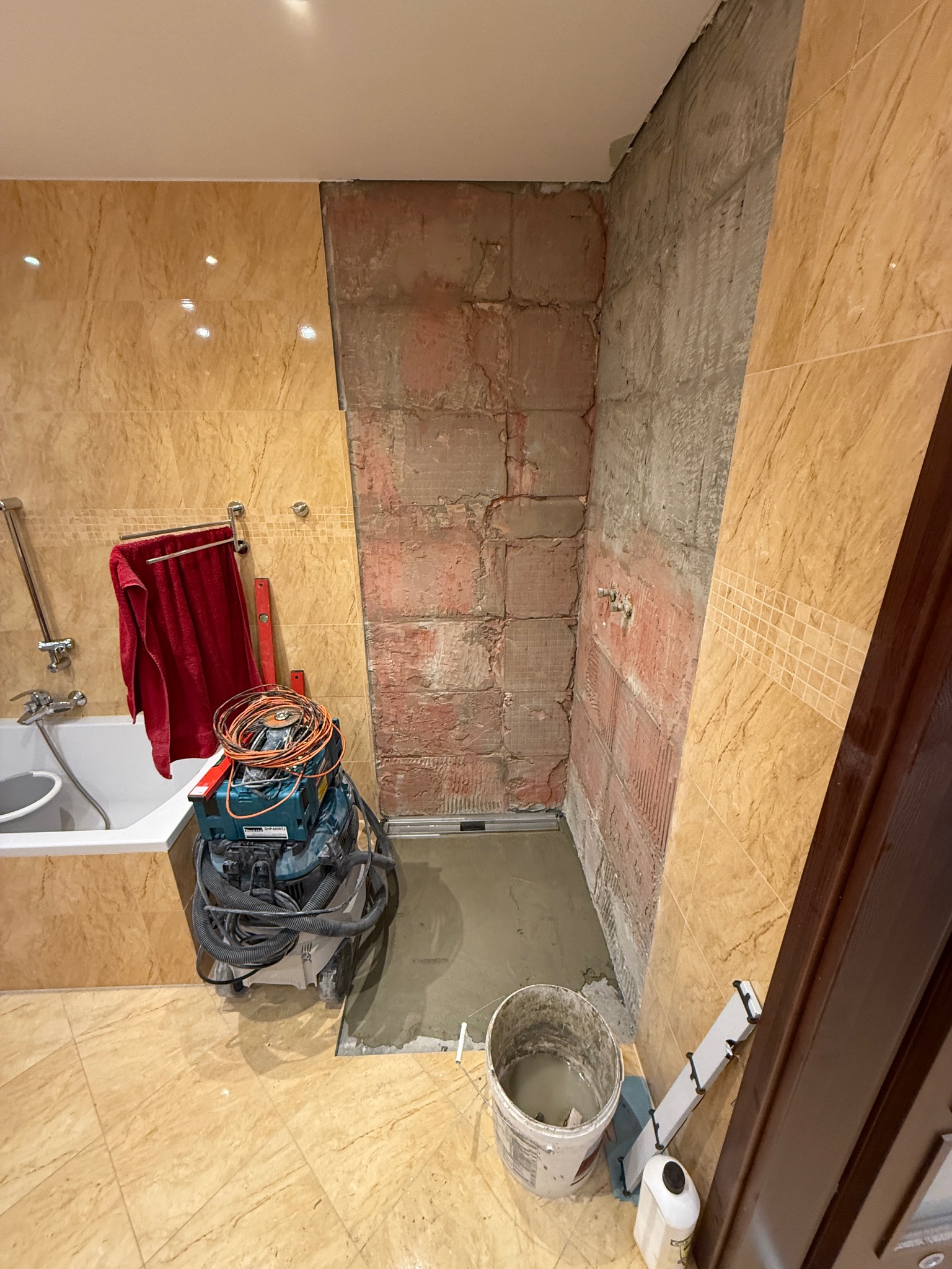 Fragment łazienki w trakcie prac. Widoczna ściana z odsłoniętą cegłą, częściowo wykończona płytkami imitującymi marmur. Na podłodze narzędzia i wiadro z zaprawą.
