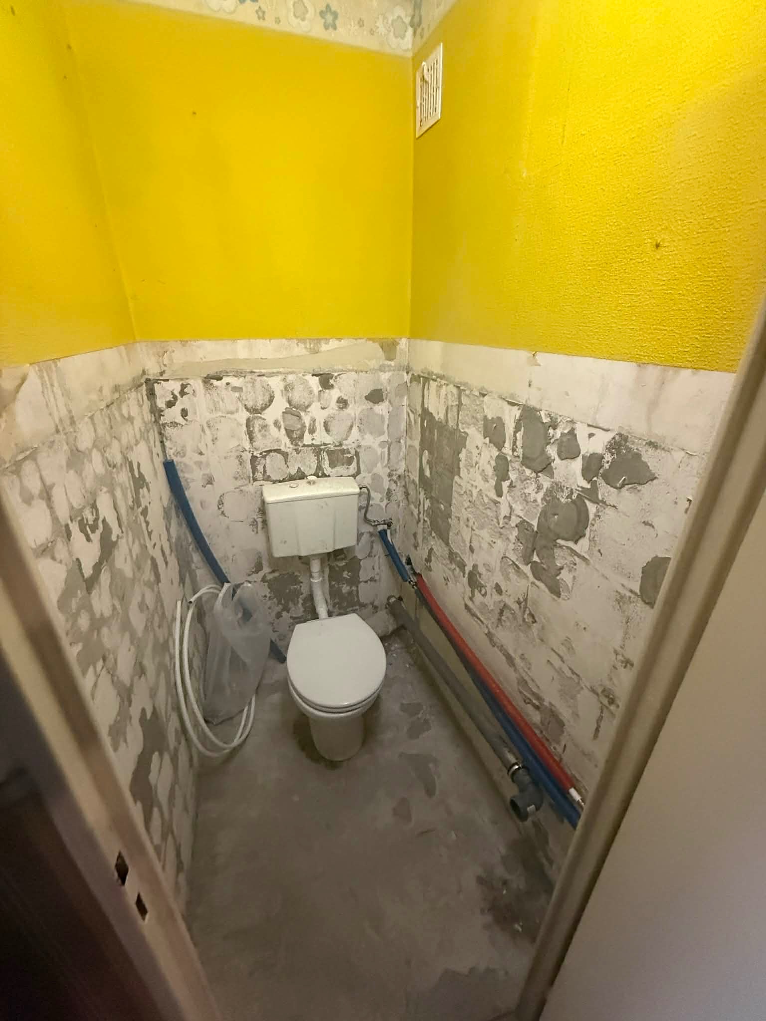 Łazienka w trakcie remontu: żółte ściany, częściowo usunięte płytki, widoczne rury instalacyjne i toaleta. Przygotowanie do dalszych prac wykończeniowych.